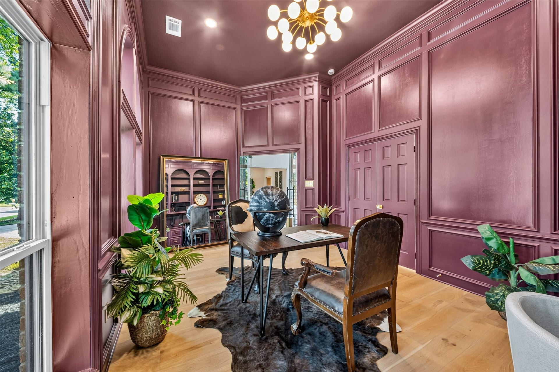 46 Pebble Cove Drive, The Woodlands, TX 77381 Elegant home office with rich burgundy paneling, warm wood floors, and abundant natural light. A sophisticated chandelier and lush greenery create a refined atmosphere.