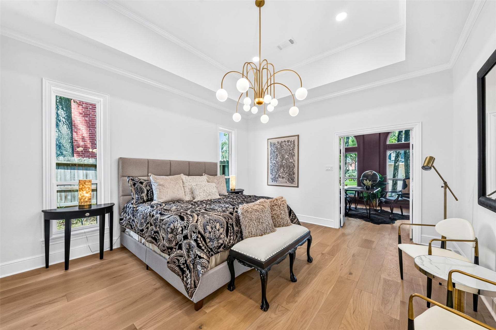 46 Pebble Cove Drive, The Woodlands, TX 77381 Elegant bedroom featuring a stylish chandelier, tray ceiling, and abundant natural light from large windows. The room seamlessly connects to a cozy sitting area, offering a perfect blend of comfort and sophistication.