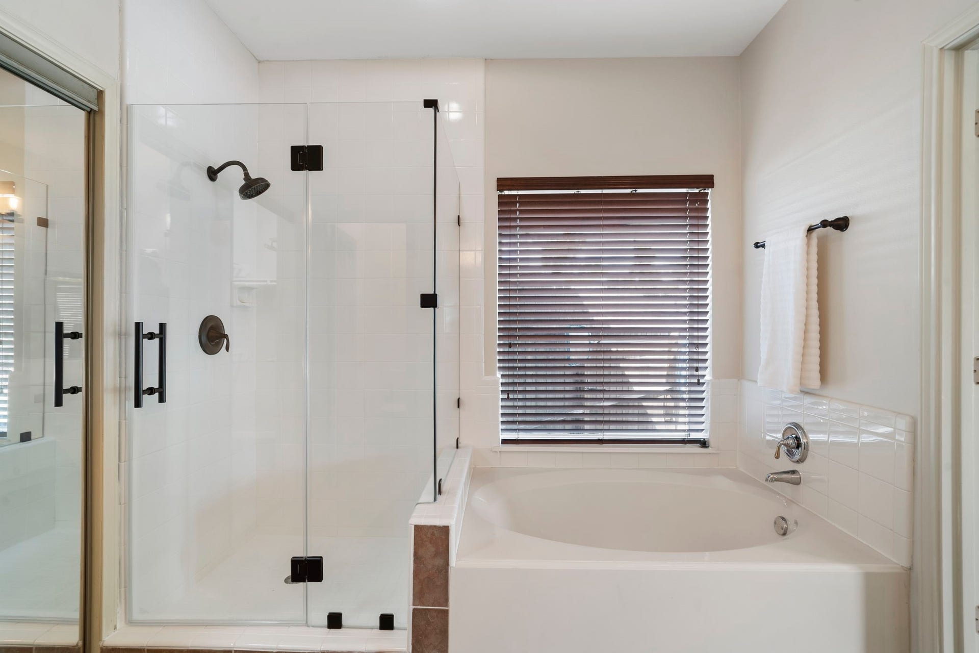 9730 Harrison Lane, Missouri City, TX 77459 Luxurious primary bath featuring a deep oval soaking tub, seamless glass-enclosed walk-in shower with dark hardware accents, white tile surround, and custom window blinds.
