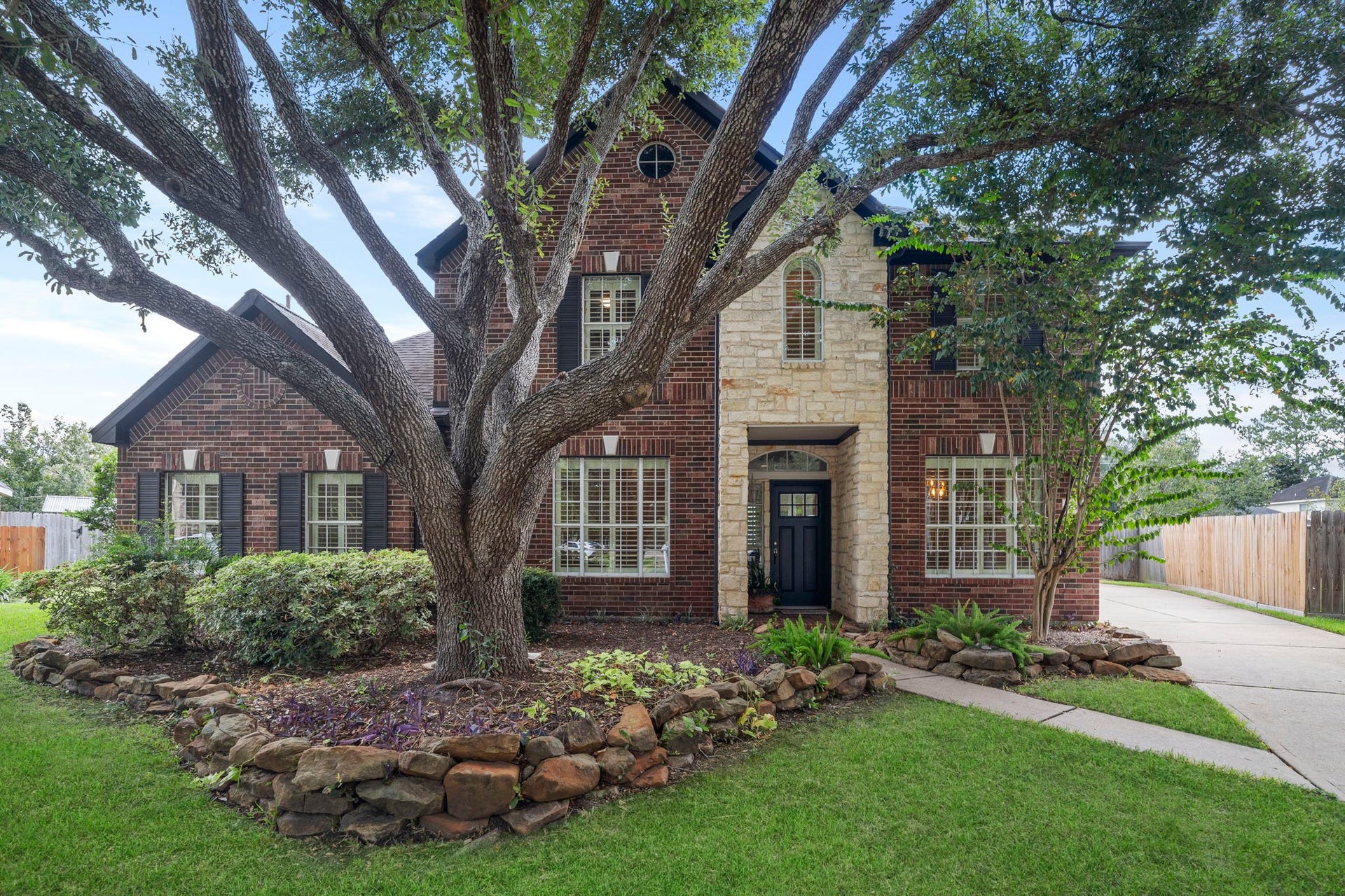 9730 Harrison Lane, Missouri City, TX 77459 Classic red brick and stone elevation framed by a dramatic mature oak shade tree, stone-bordered flower beds, and custom window shutters.