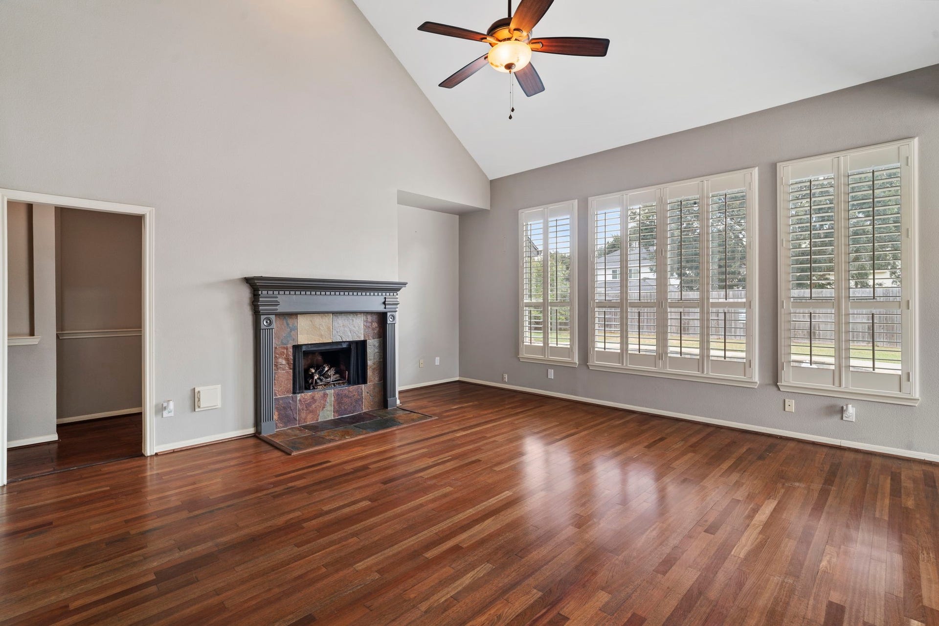 9730 Harrison Lane, Missouri City, TX 77459 Expansive main family room featuring dramatic vaulted ceilings, wood-look flooring, ceiling fan, slate-surround gas fireplace, and a wall of windows framed by custom plantation shutters.