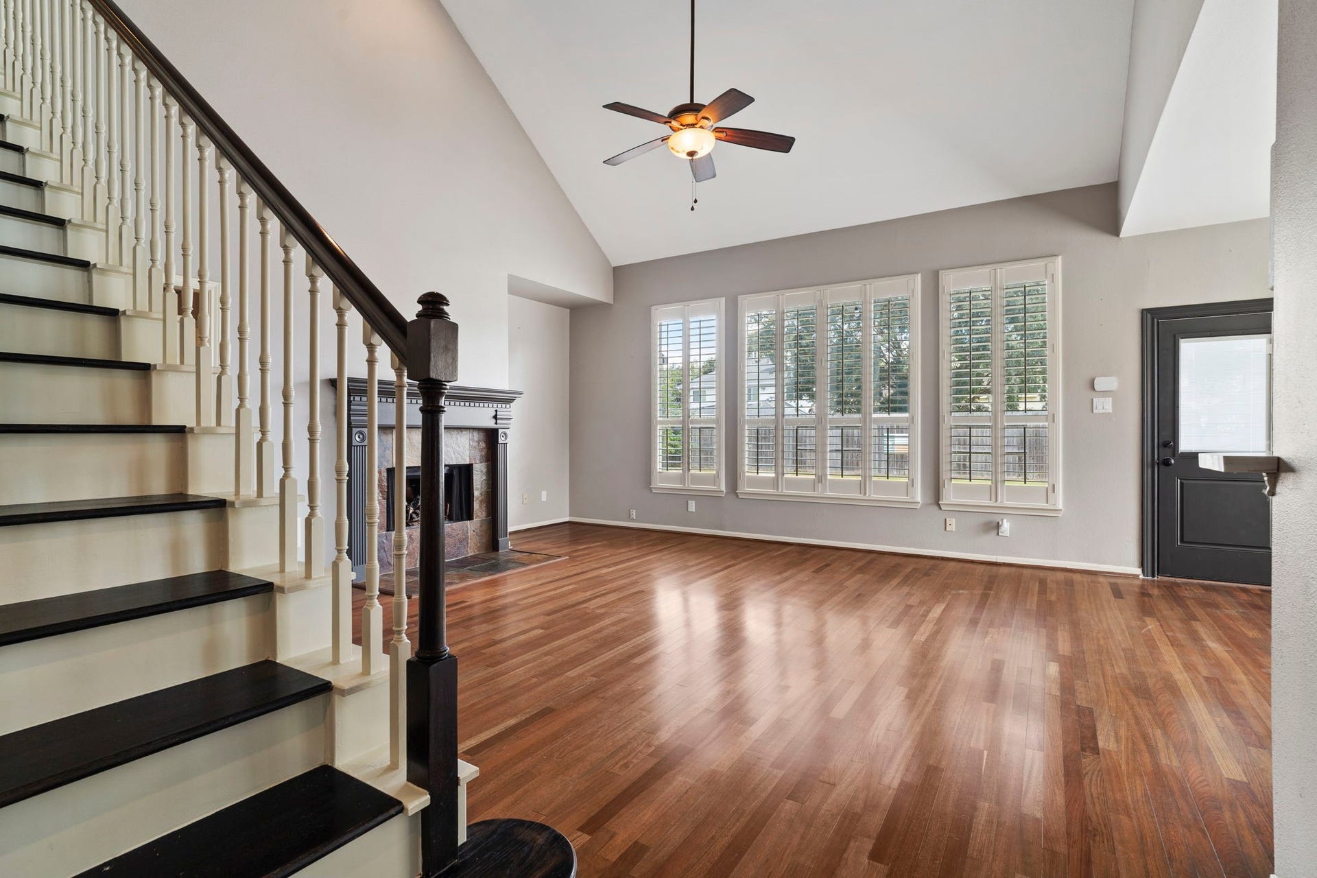 9730 Harrison Lane, Missouri City, TX 77459 Soaring two-story living room showcasing a corner stone gas fireplace, rich wood-look floors, tall shuttered windows, ceiling fan, and open staircase access.