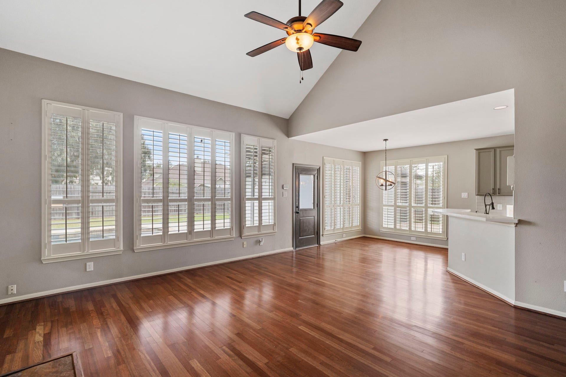 9730 Harrison Lane, Missouri City, TX 77459 Vaulted main living area showing the high wall of shuttered windows, rich wood-look floors, direct exterior door access, and open flow into the breakfast room and kitchen counter.