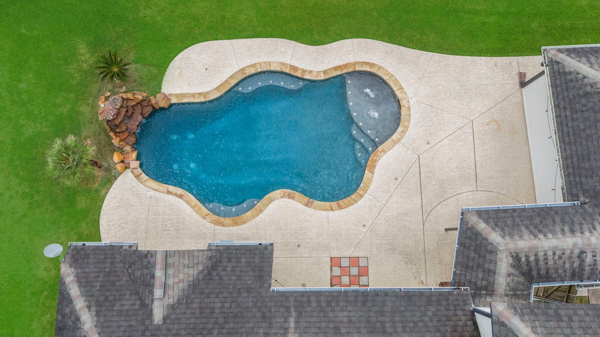 9730 Harrison Lane, Missouri City, TX 77459 Overhead drone capture showcasing the freeform swimming pool layout, rock waterfall feature, tanning ledge, cool-deck surround patio, and lush green grass.