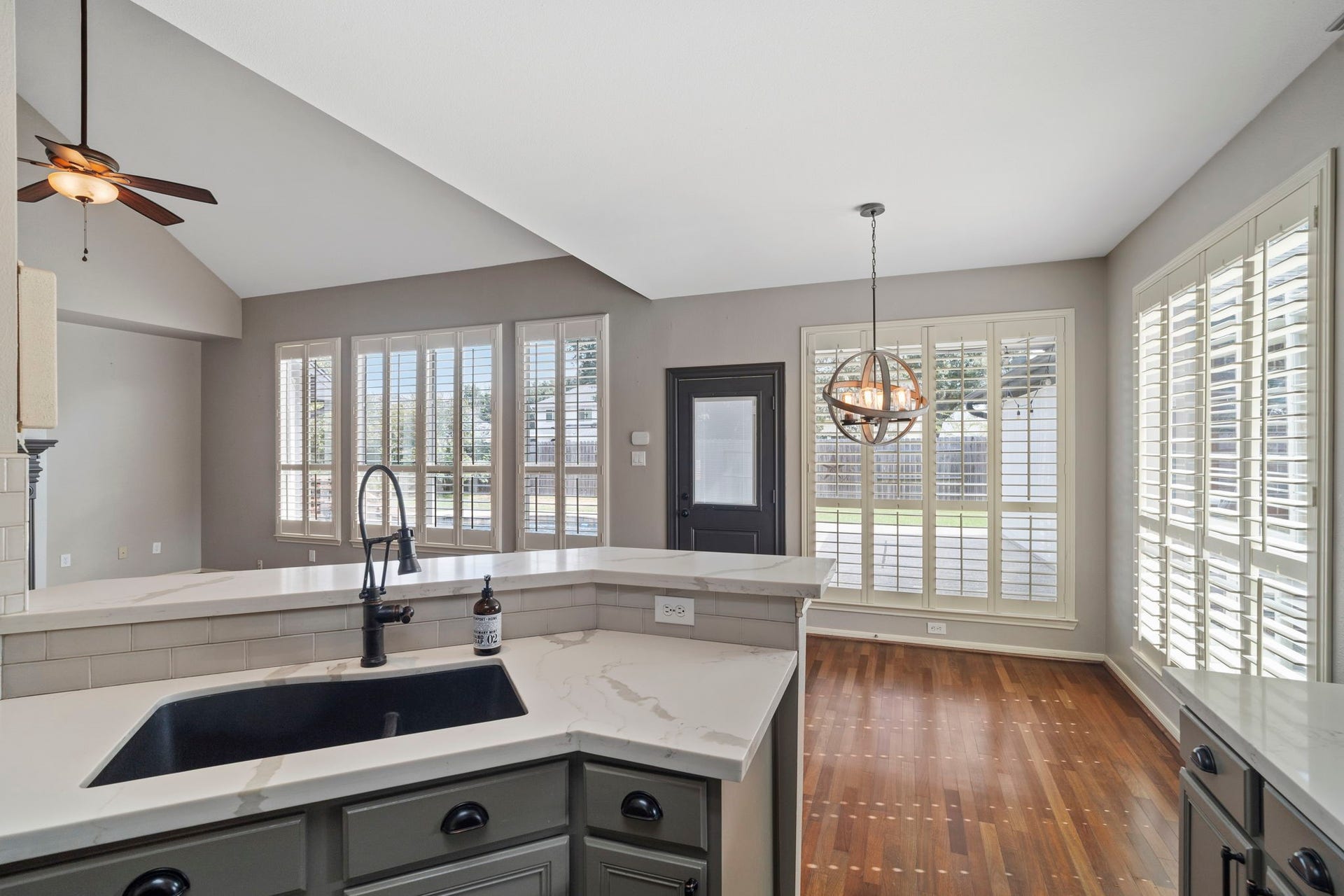 9730 Harrison Lane, Missouri City, TX 77459 Kitchen sink peninsula view looking out toward the sunlit breakfast nook with custom plantation shutters, dark undermount sink with gooseneck faucet, quartz countertops, and soft sage cabinetry.