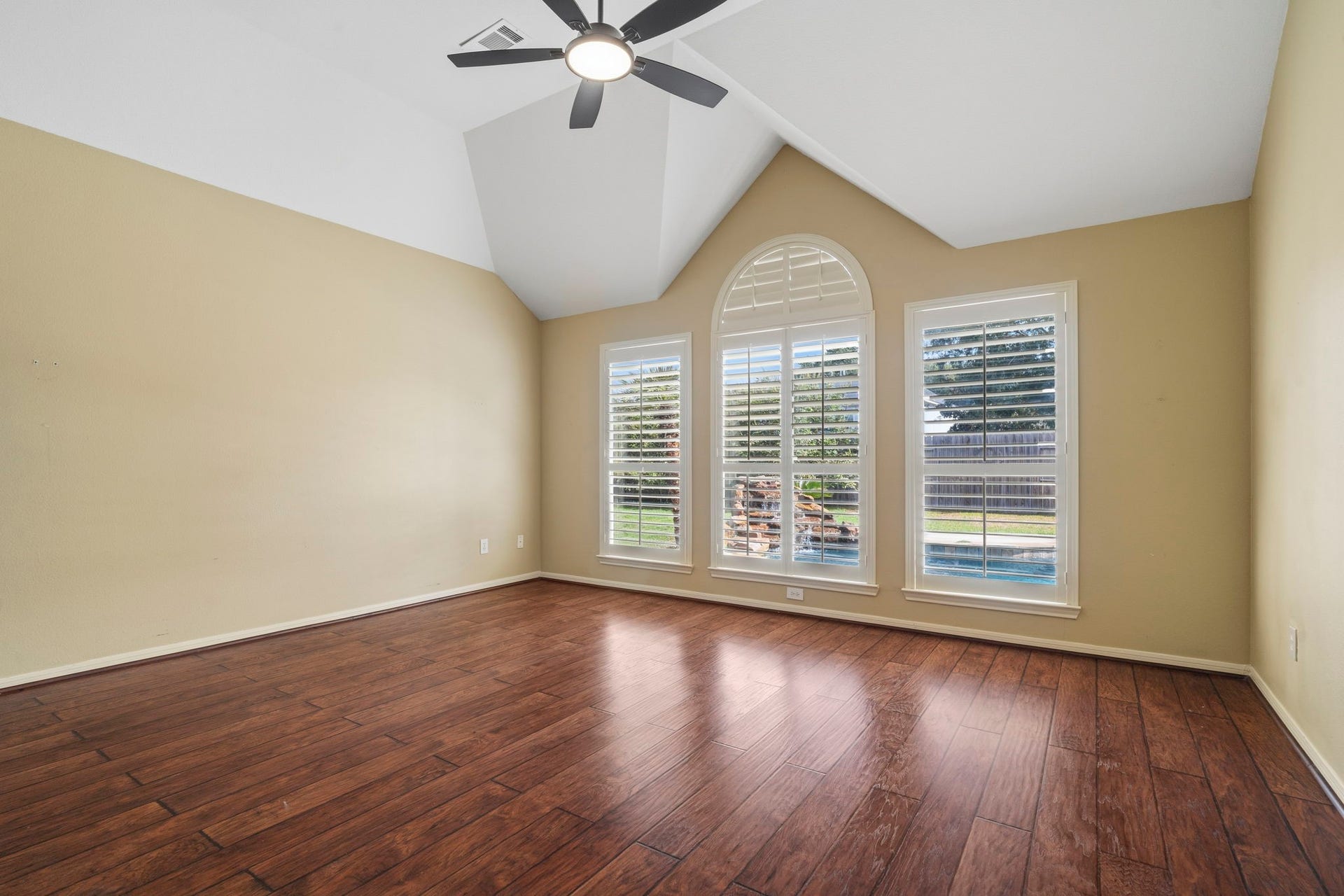 9730 Harrison Lane, Missouri City, TX 77459 Primary bedroom featuring soaring vaulted ceilings, wood-look floors, ceiling fan, and large windows with custom arched plantation shutters looking out to the pool area.