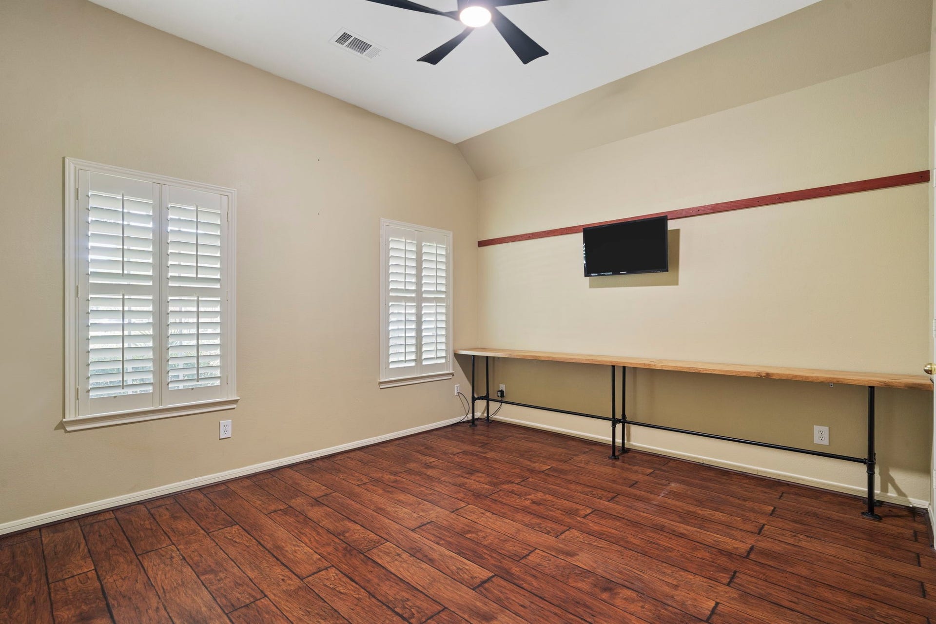 9730 Harrison Lane, Missouri City, TX 77459 Main-floor office / study showing custom plantation window shutters, dark wood-look flooring, high ceilings with ceiling fan, wall-mounted television setup, and an integrated industrial pipe-and-wood built-in long desk station.