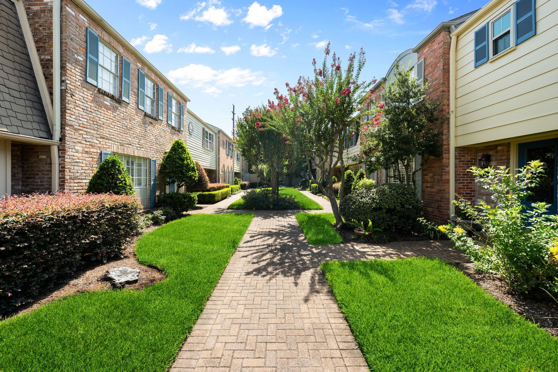 5819 Valley Forge Drive, #91, Houston, TX 77057 Charming townhomes with classic brick facades line a beautifully landscaped walkway, featuring lush greenery and mature trees under a bright blue sky.