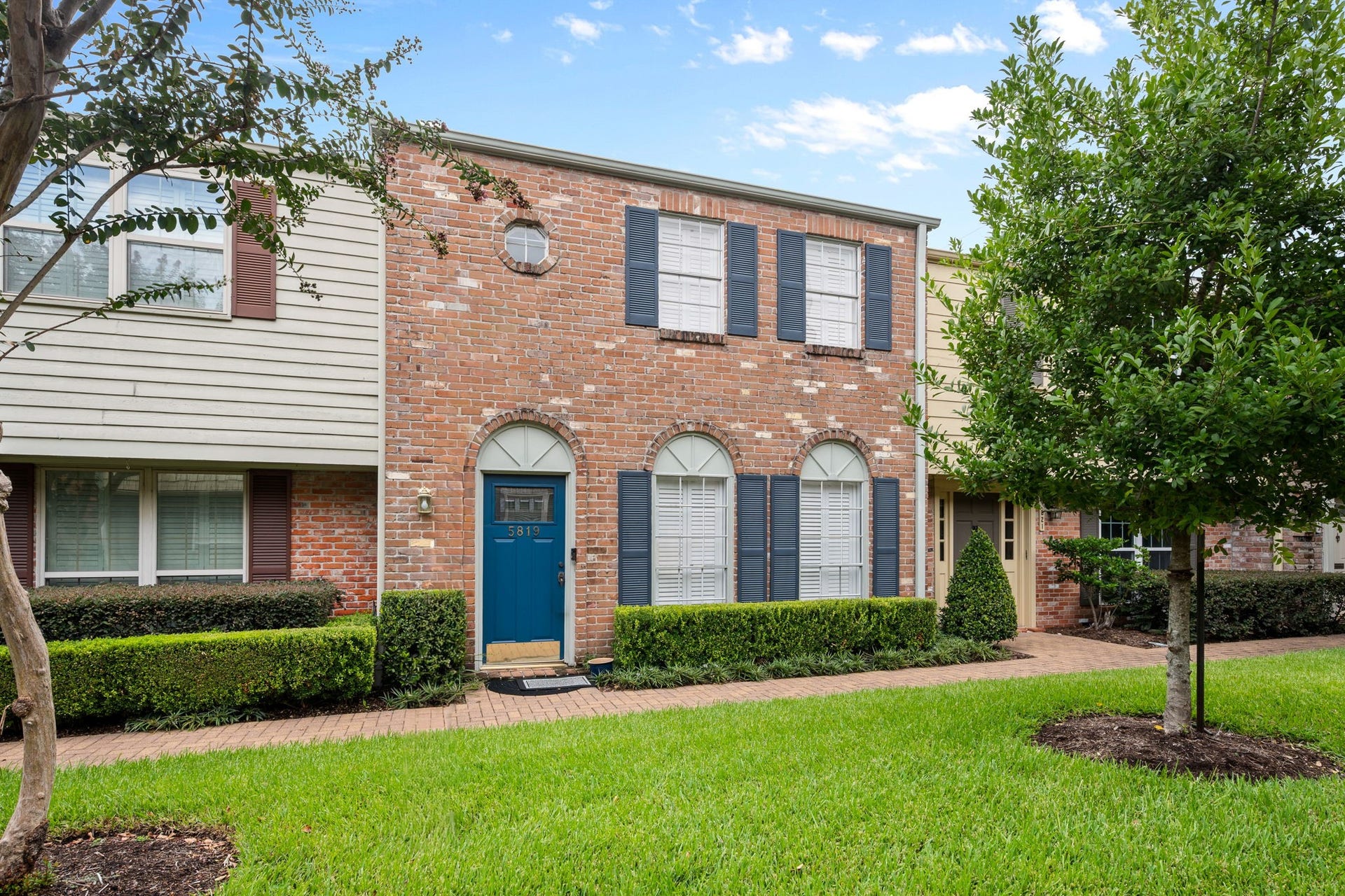 5819 Valley Forge Drive, #91, Houston, TX 77057 Charming brick townhouse with elegant shutters, arched windows, and a vibrant blue front door, nestled in a well-maintained neighborhood with lush greenery.