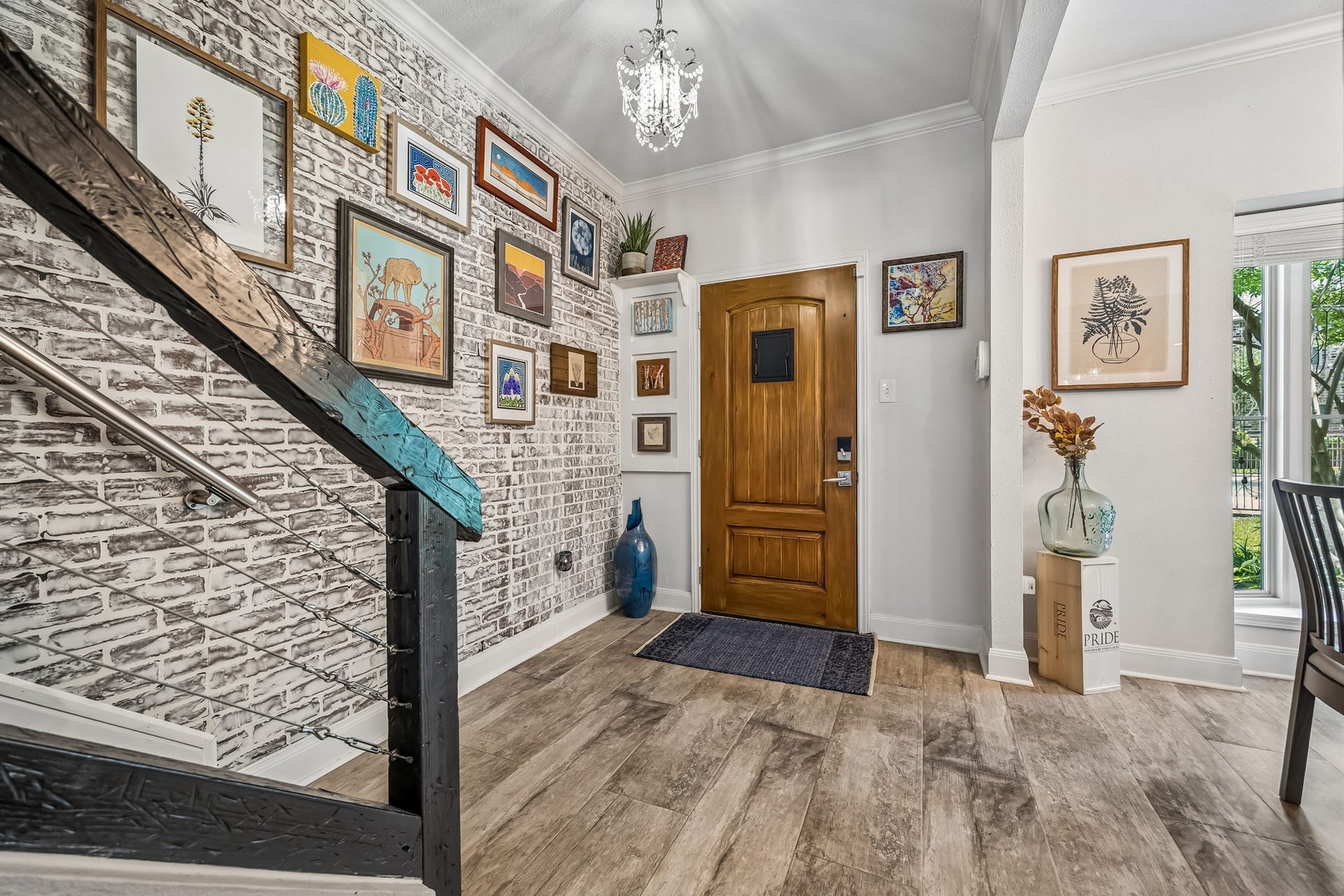 14719 Barryknoll Lane, #51, Houston, TX 77079 A warm wood front door opens to a stylish entry with wood-look flooring, crown molding, and a statement chandelier. The brick accent wall and stair detail add character, while nearby windows bring in a view of the surrounding greenery.