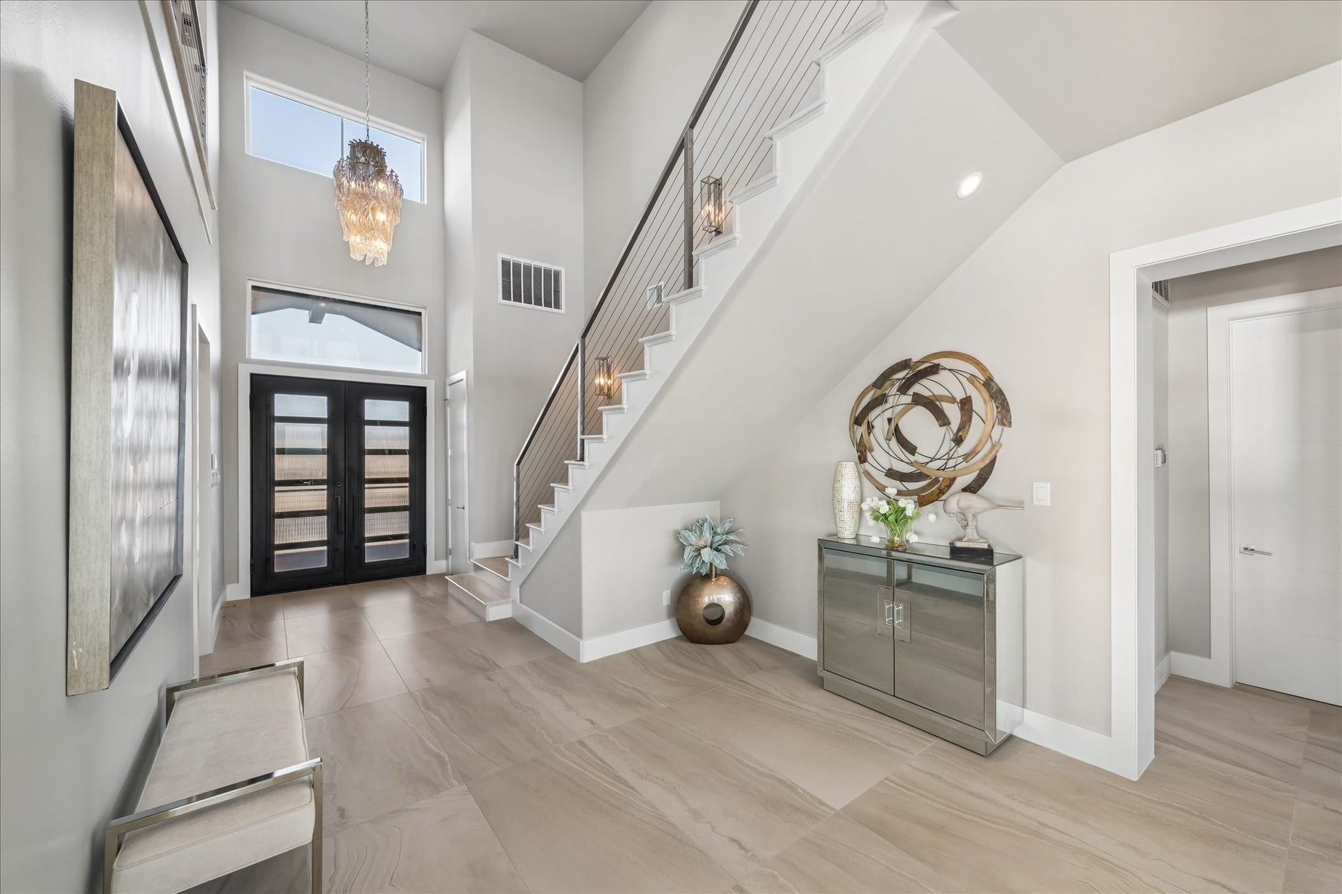1 Water Key, Hitchcock, TX 77563 A grand, foyer showcases soaring ceilings, abundant natural light, and modern double doors with glass lites and a transom window. A staircase with wood treads and cable railing anchors the space, complemented by a dramatic chandelier and sleek Eleganza tile flooring