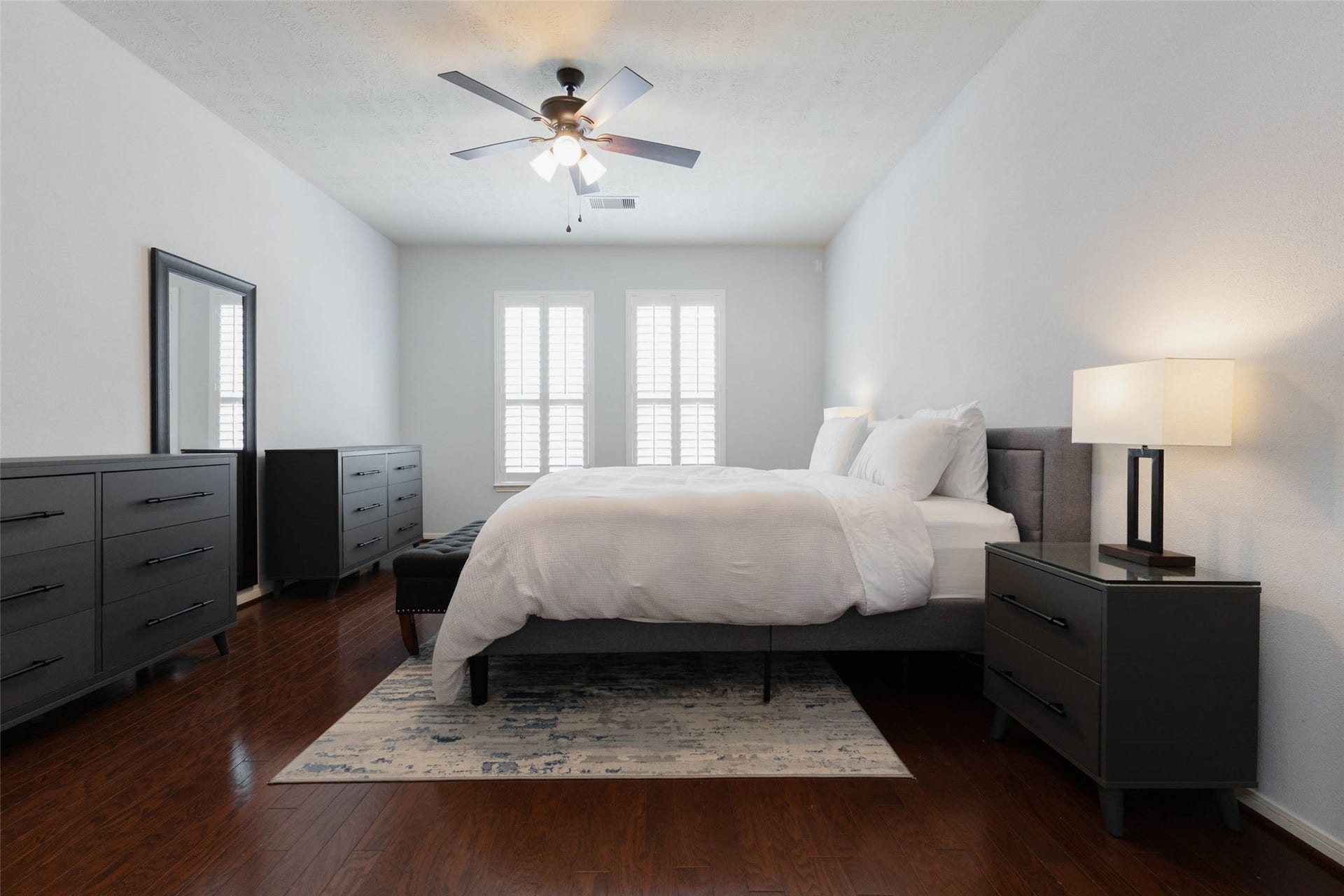 Retreat to the spacious primary suite, where rich wood-look flooring, soft neutral tones, and generous proportions create a comfortable private escape. A pair of windows dressed with plantation shutters welcomes natural light, while a ceiling fan adds everyday comfort.