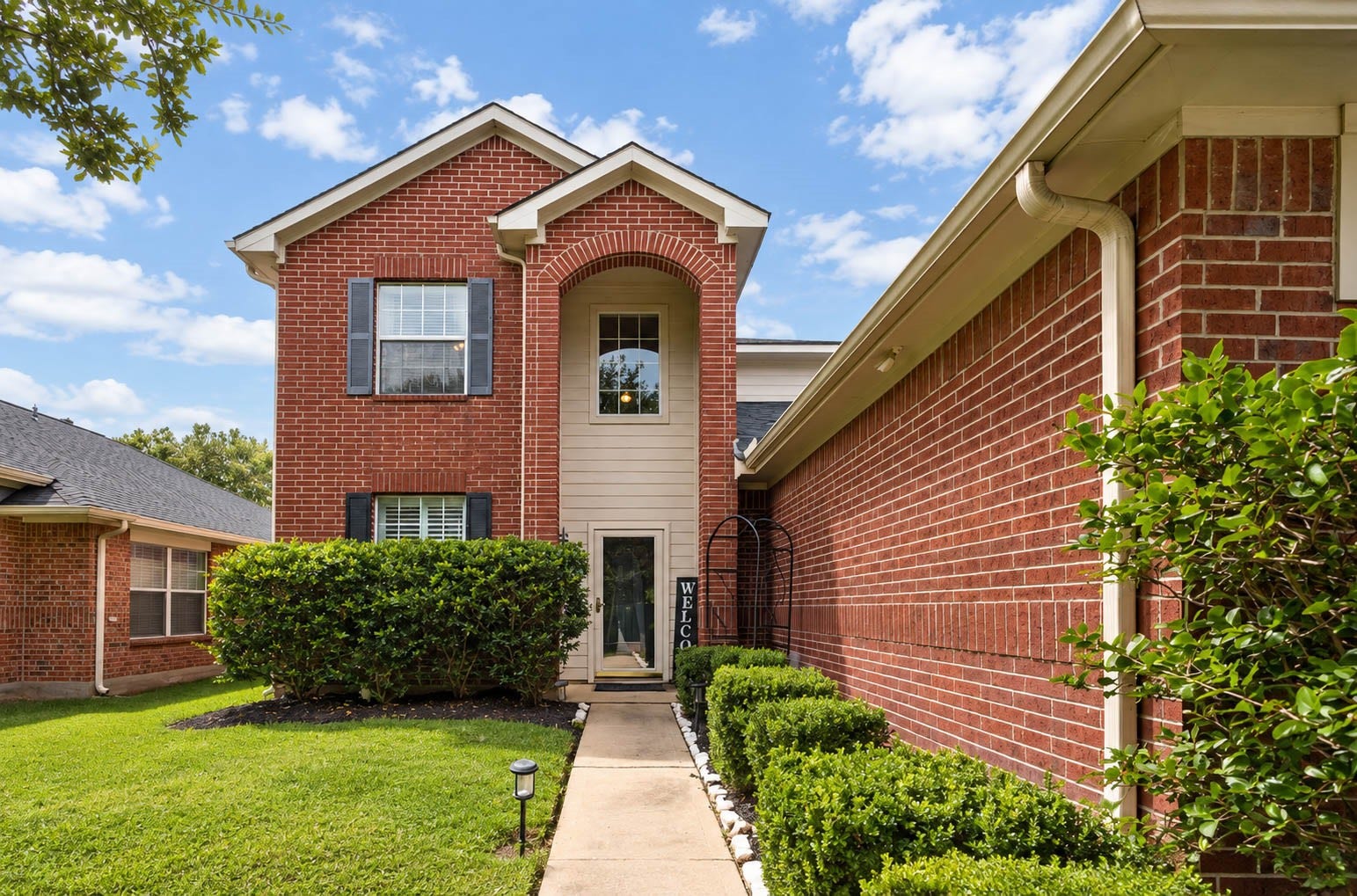 A closer look at the front elevation highlights traditional brick architecture, an arched entry, and neatly maintained landscaping. Mature hedges line the walkway leading to the sheltered front door.