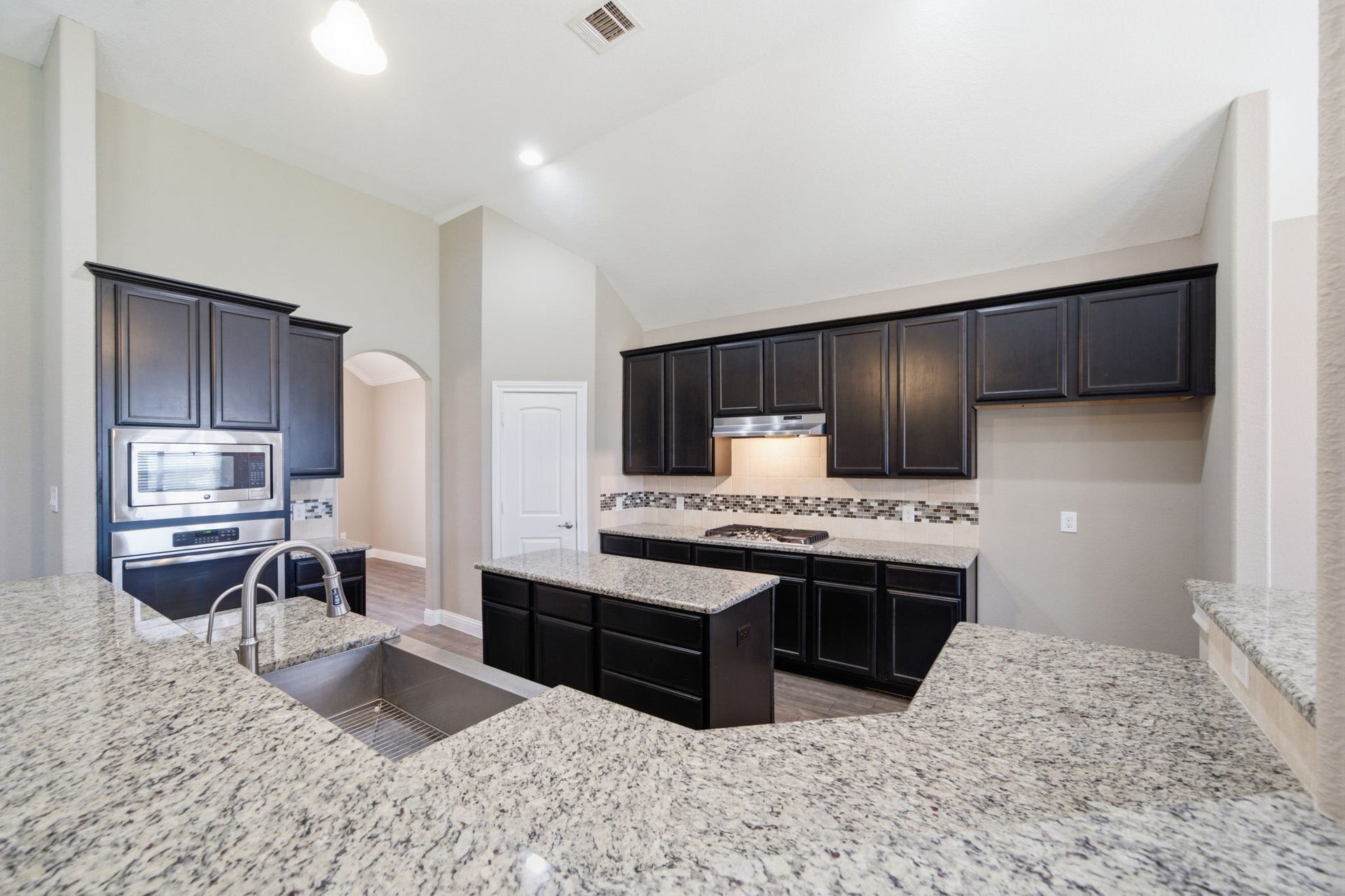 18818 Wichita Trail, Magnolia, TX 77355 Polished granite countertops sweep across a breakfast bar with an undermount stainless steel sink, facing a central island and a gas cooktop. High vaulted ceilings, dark cabinetry, and a mosaic tile backsplash highlight this functional open-concept culinary space.