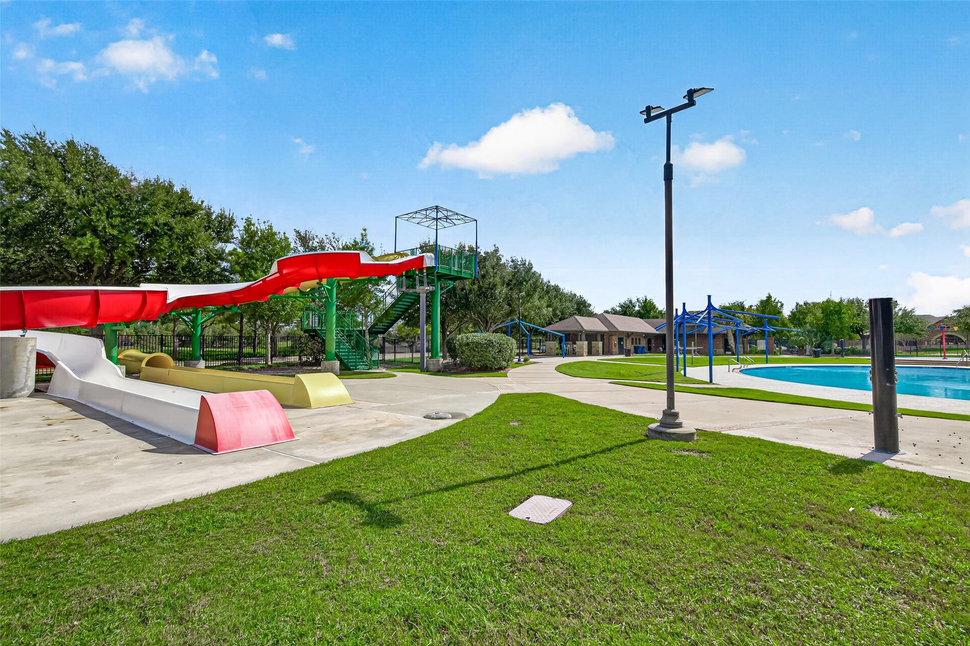 Experience true water-park thrills right in your own community with this exciting water slide attraction! Towering over the pool grounds, a multi-story green stairs structure leads up to long, twisting open and enclosed slides in vibrant red, white, and yellow tones that plunge into smooth landing chutes below. Wide concrete pathways connect the slide exit area seamlessly to the nearby resort pool, shaded lounge spaces, and lush green lawns.