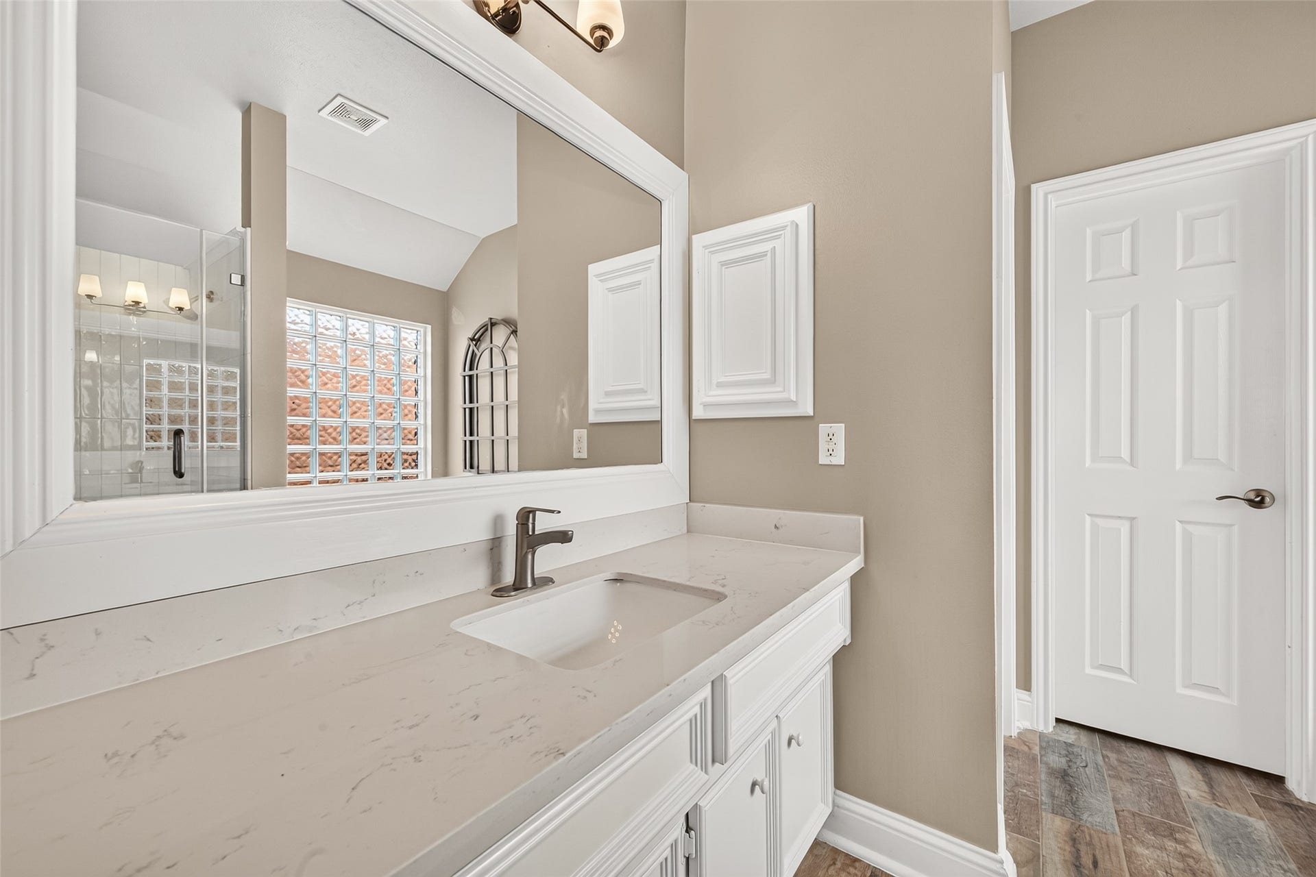 Indulge in spa-like comfort in this beautifully updated primary bathroom vanity area, crafted to make your daily routine feel like a luxury experience. The expansive white vanity features a sleek quartz countertop with subtle marble-look veining, an undermount rectangular sink, and modern dark bronze hardware. Framed by a large white molding mirror, the vanity reflects the soft ambient light and high ceiling details of the room.