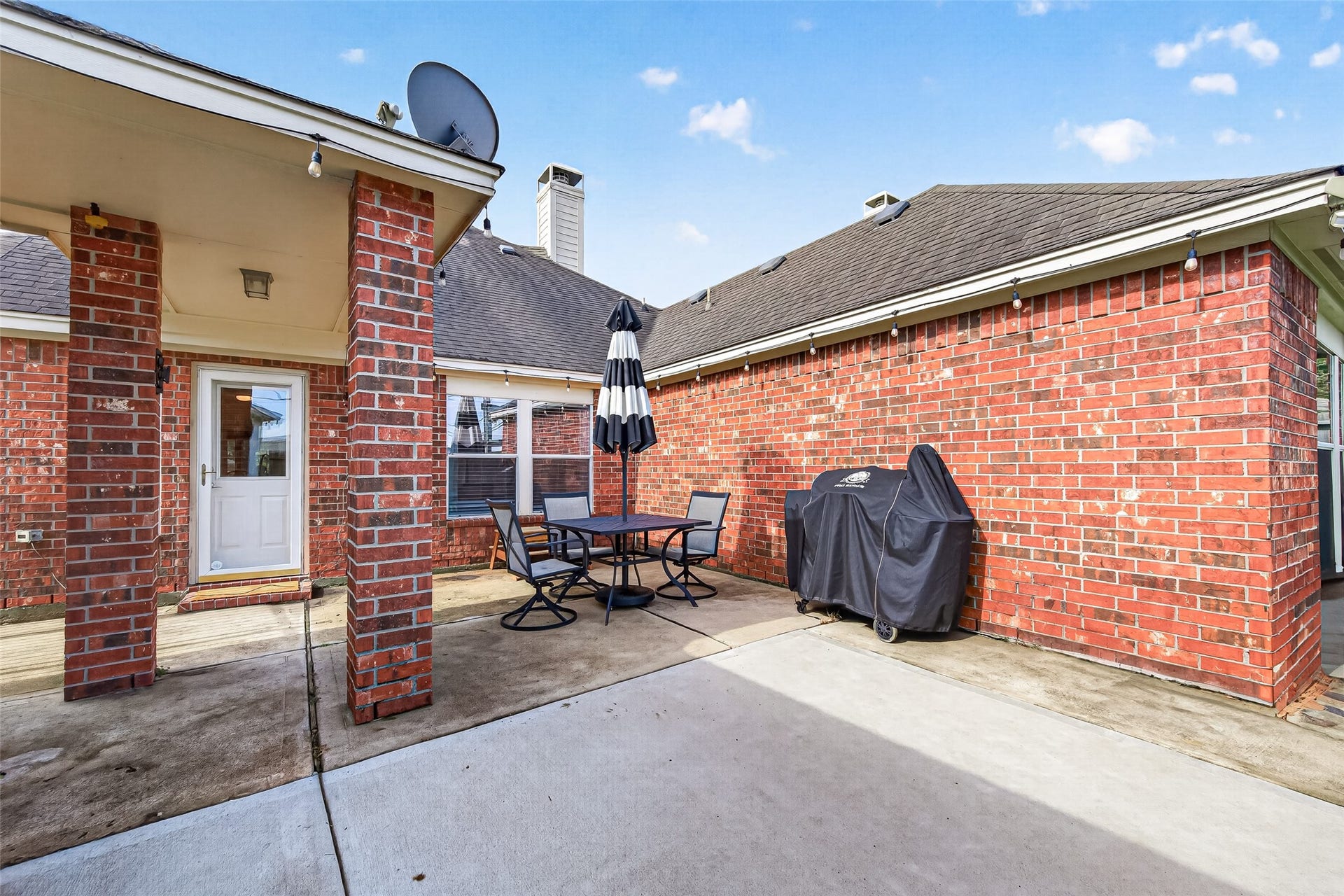 Step outside to your private outdoor sanctuary, designed for effortless relaxation and weekend entertaining. This spacious concrete patio features a covered porch area anchored by sturdy red brick support pillars and ambient string lighting strung along the roofline.