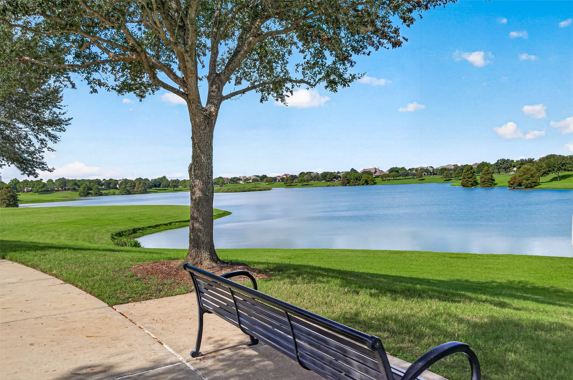 Showcasing the serene natural beauty of the community, this picturesque lakefront setting offers a tranquil escape for outdoor relaxation and quiet contemplation. A dark metal park bench sits beneath the generous shade of a mature oak tree, positioned alongside a smooth concrete walking path.