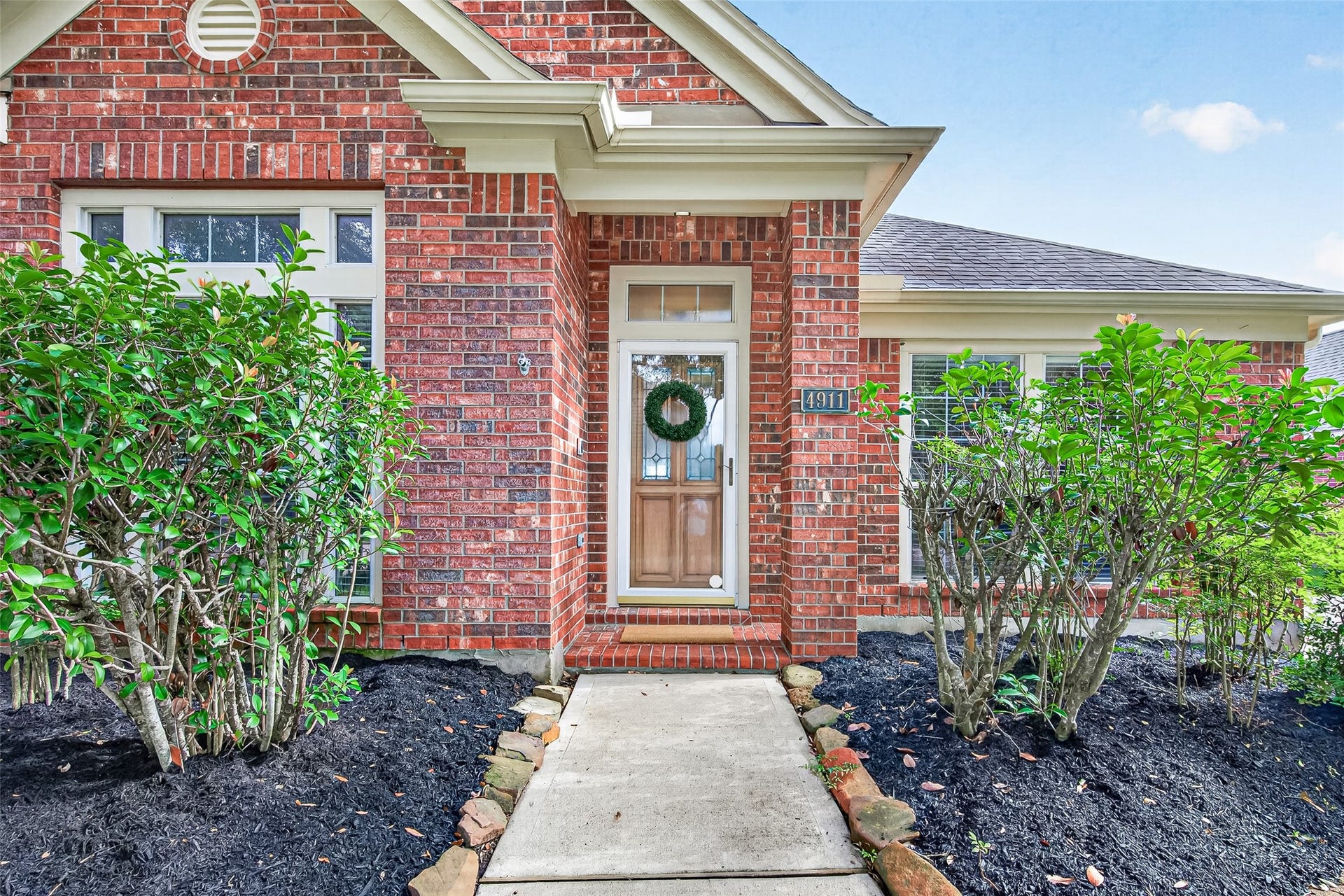 Raised, stacked-stone borders encircle the bases of the trees, filled with dark mulch, while matching mulched garden beds lined with neatly trimmed green bushes run along the entire base of the front elevation.