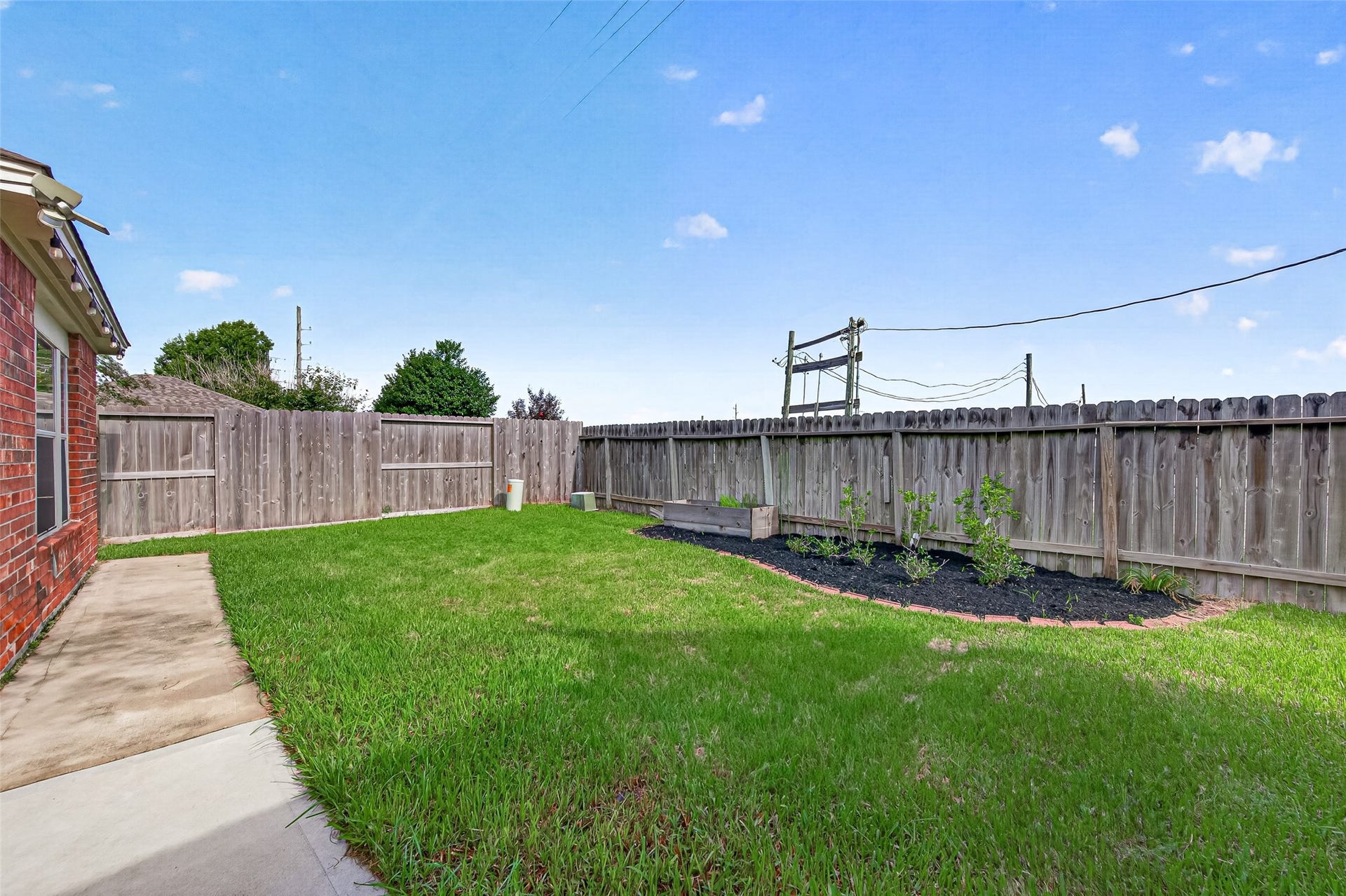 This fully fenced backyard offers a private, low-maintenance outdoor retreat ideal for pets, play, and gardening enthusiasts. A lush green lawn stretches along the rear elevation, highlighted by a stone-bordered garden bed filled with dark mulch and a raised planter box perfect for growing fresh herbs or vegetables.