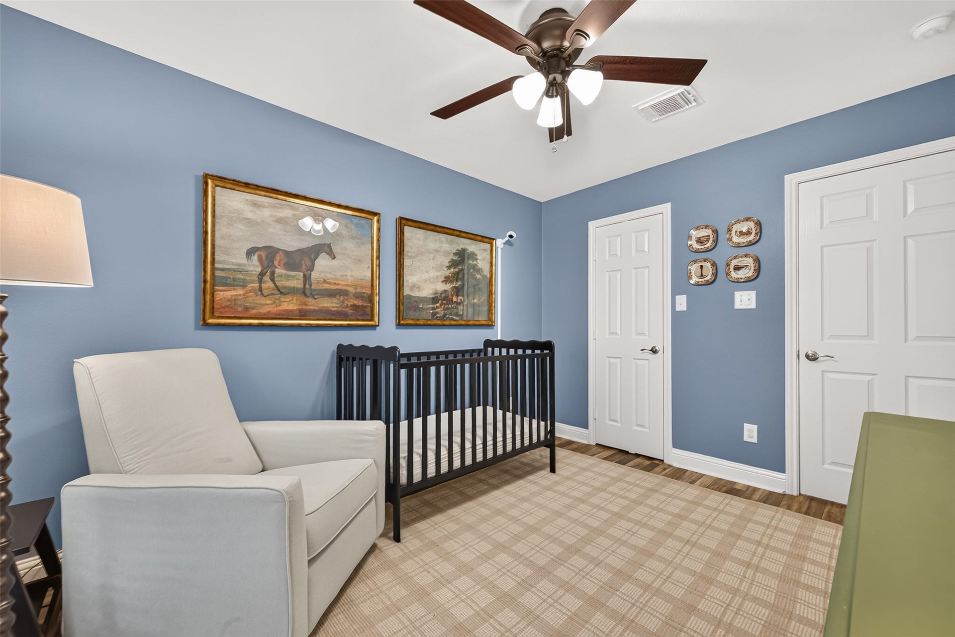 Soft blue-grey painted walls are complemented by crisp white baseboards and rich multi-tone wood-look plank flooring. A large window fitted with horizontal blinds streams in pleasant natural light while overlooking lush exterior greenery.