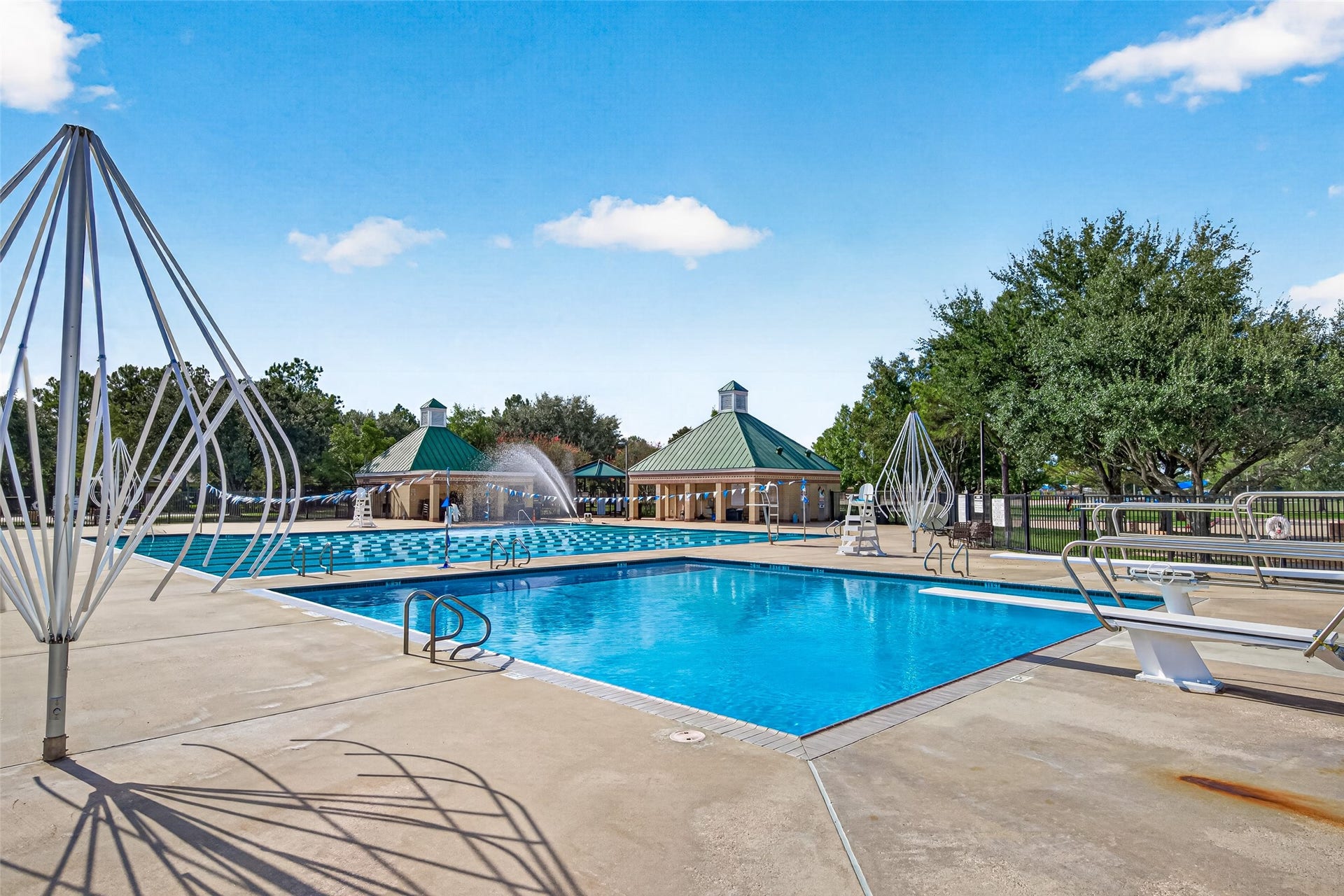 Dive into ultimate relaxation and recreation at this expansive community aquatic center! Designed to accommodate both competitive swimmers and leisure seekers, the facility features a large lap pool equipped with designated swimming lanes, water fountains, and lifeguard stands, alongside a dedicated diving pool complete with dual diving boards. Surrounding the sparkling clear blue water is a broad concrete pool deck with shade structures, green-roofed bathhouses, and serene views of lush surrounding trees u