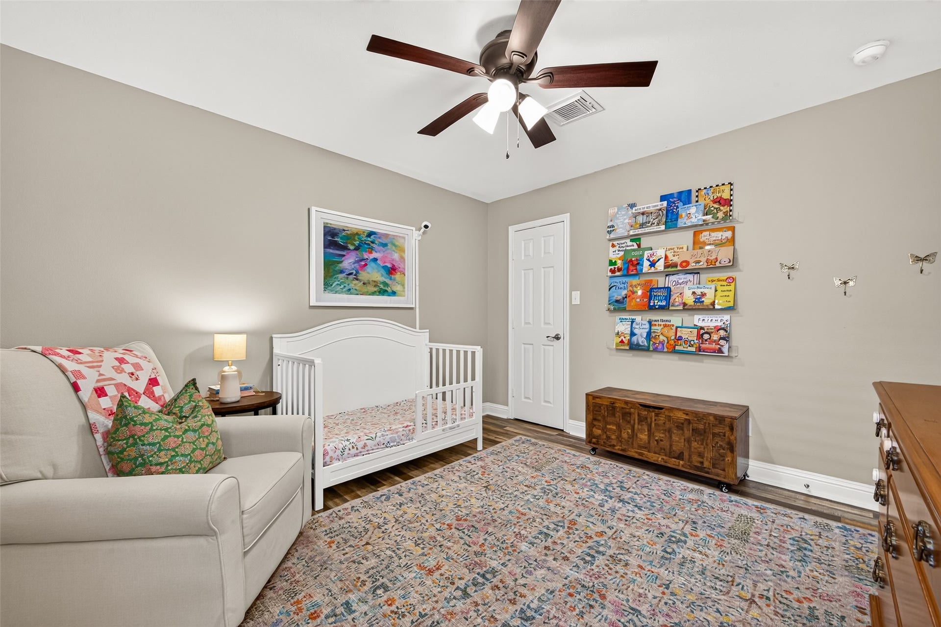 A large window outfitted with horizontal blinds and framed by full-length curtains brings in abundant natural light while framing views of the exterior brick detail. High ceilings are topped with a traditional wood-blade ceiling fan featuring an integrated light fixture for continuous comfort.