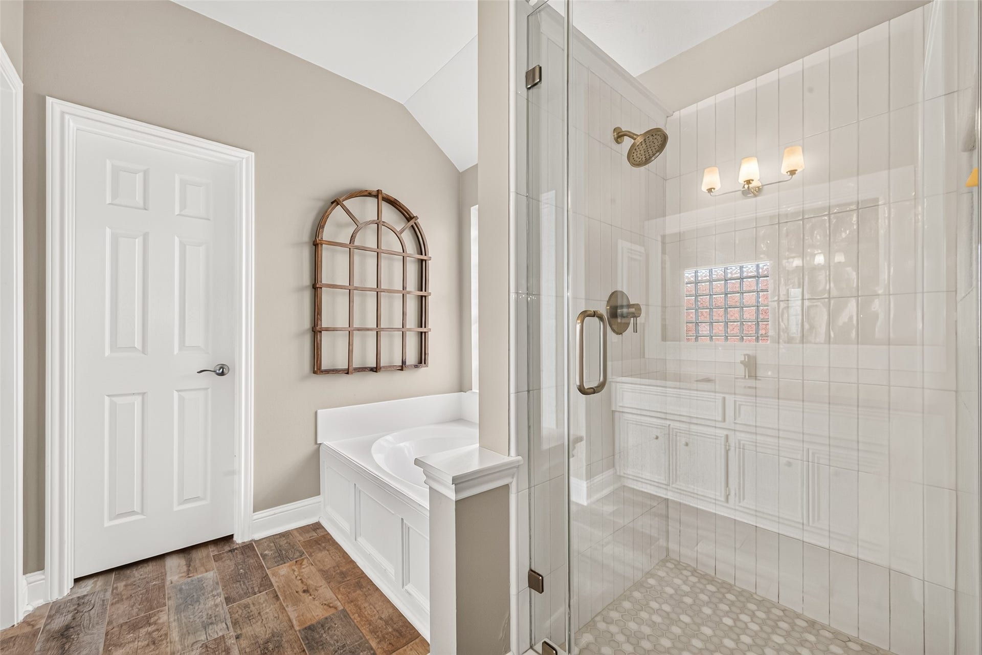 Designed as a tranquil home spa, this primary bathroom layout offers both a deep soaking tub and a walk-in glass shower.  Adjacent to the tub, the seamless frameless glass shower reveals clean white vertical subway tile walls, a classic hexagon tile floor, and elegant brushed bronze shower fixtures.