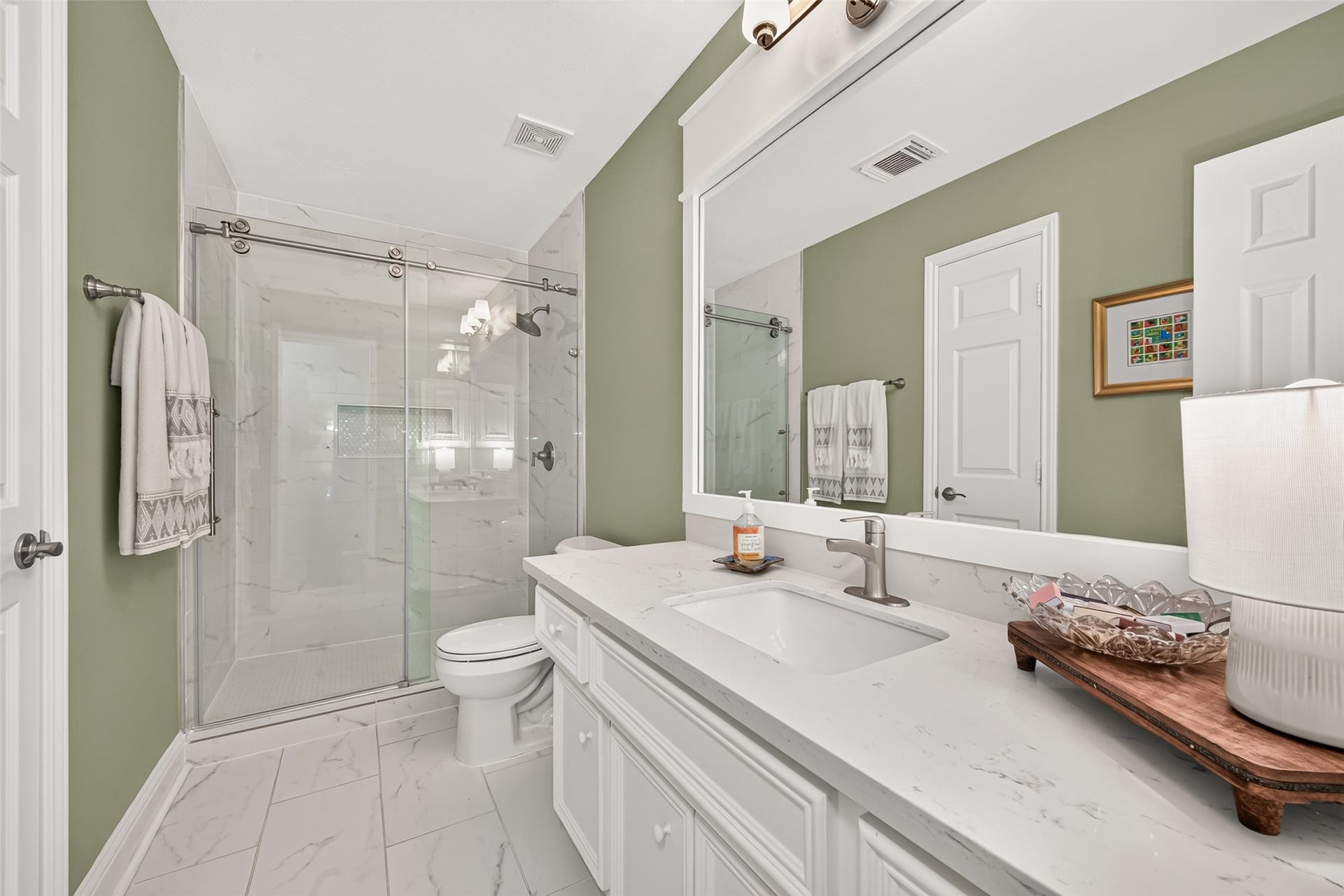 This beautifully updated full bathroom pairs sleek modern finishes with a serene sage-green color palette. An expansive white vanity features a quartz countertop with subtle marble-style veining, an undermount rectangular sink, a brushed nickel faucet, and a large white-framed mirror overhead. The walk-in shower is enclosed by frameless sliding glass doors with industrial-style exposed rollers, highlighting floor-to-ceiling marble-patterned tile, an integrated accent niche, and brushed nickel hardware.