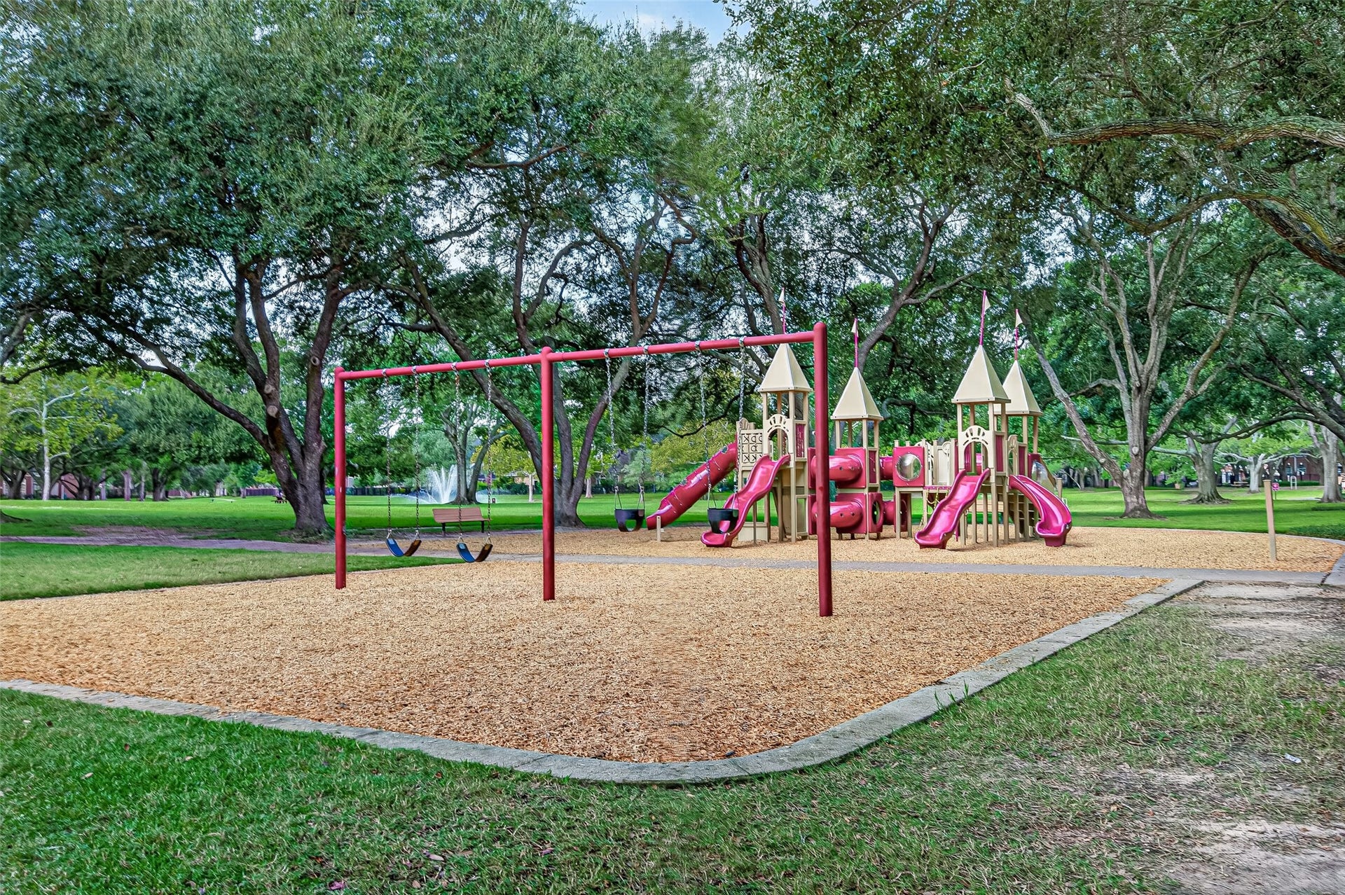 Nestled under a breathtaking canopy of mature, sprawling oak trees, this fairy-tale playground offers an enchanting setting for children to play and explore. The expansive play area features a red swing set frame with both belt and bucket toddler swings, alongside a castle-themed play structure complete with turrets, multiple slides, and climbing tunnels.