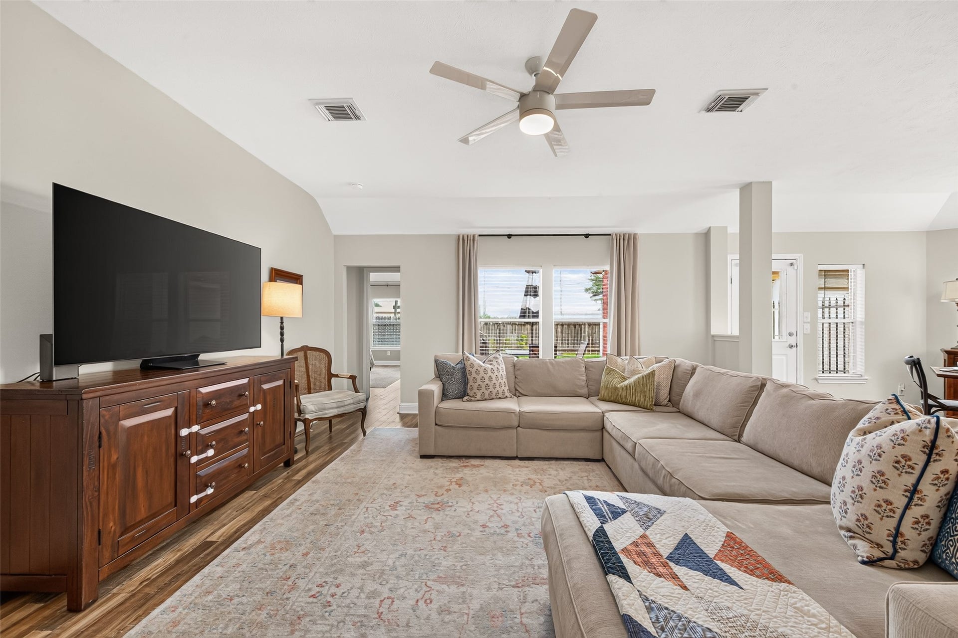 A wall of large windows frames views of the side yard while filling the room with abundant natural light. The generous floor plan comfortably accommodates a large sectional sofa, media console, and accent seating, making this inviting space the perfect gathering spot for family and guests alike.