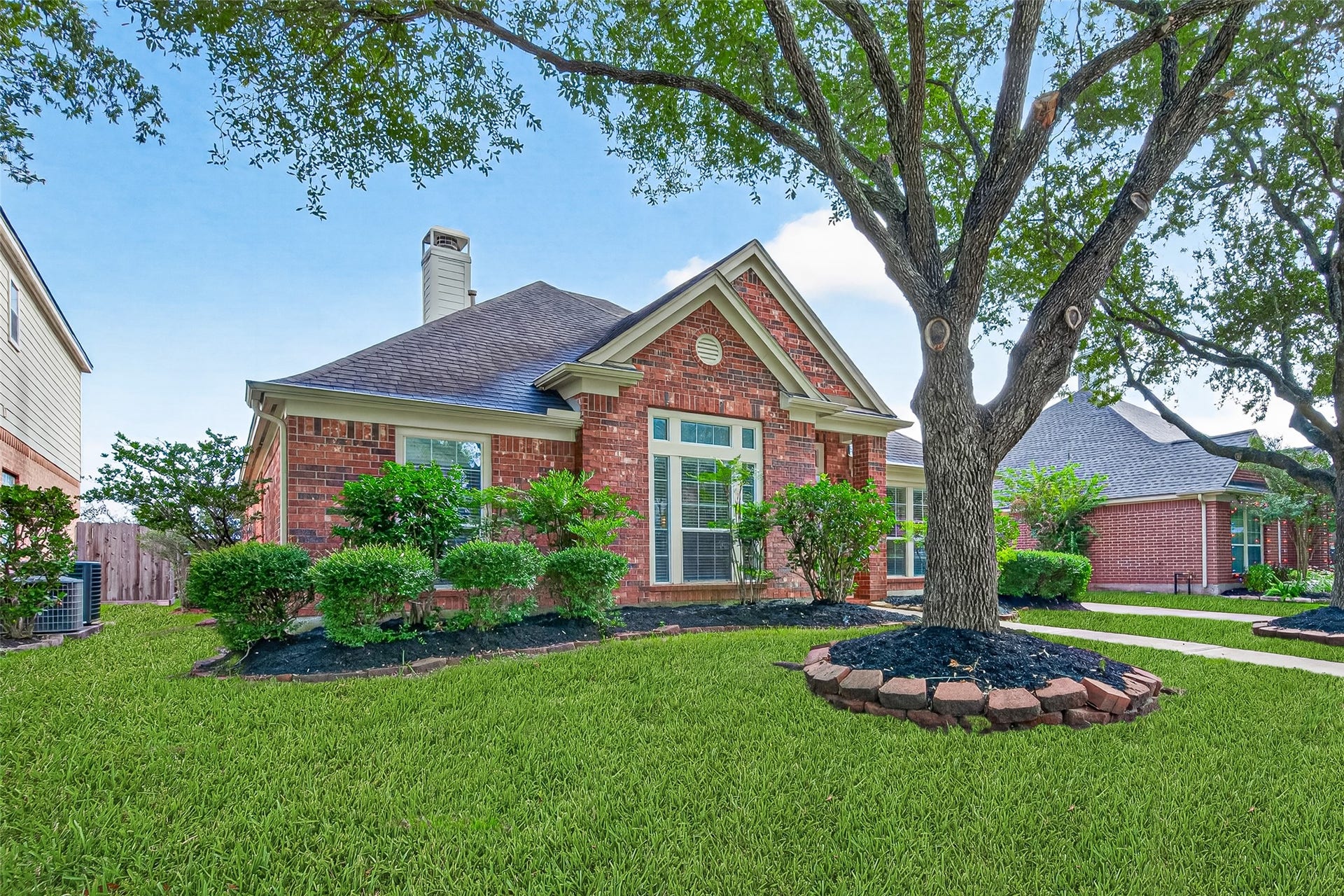 Framing the structure on both sides are two large, mature oak trees whose wide branches create a shady canopy over the yard.