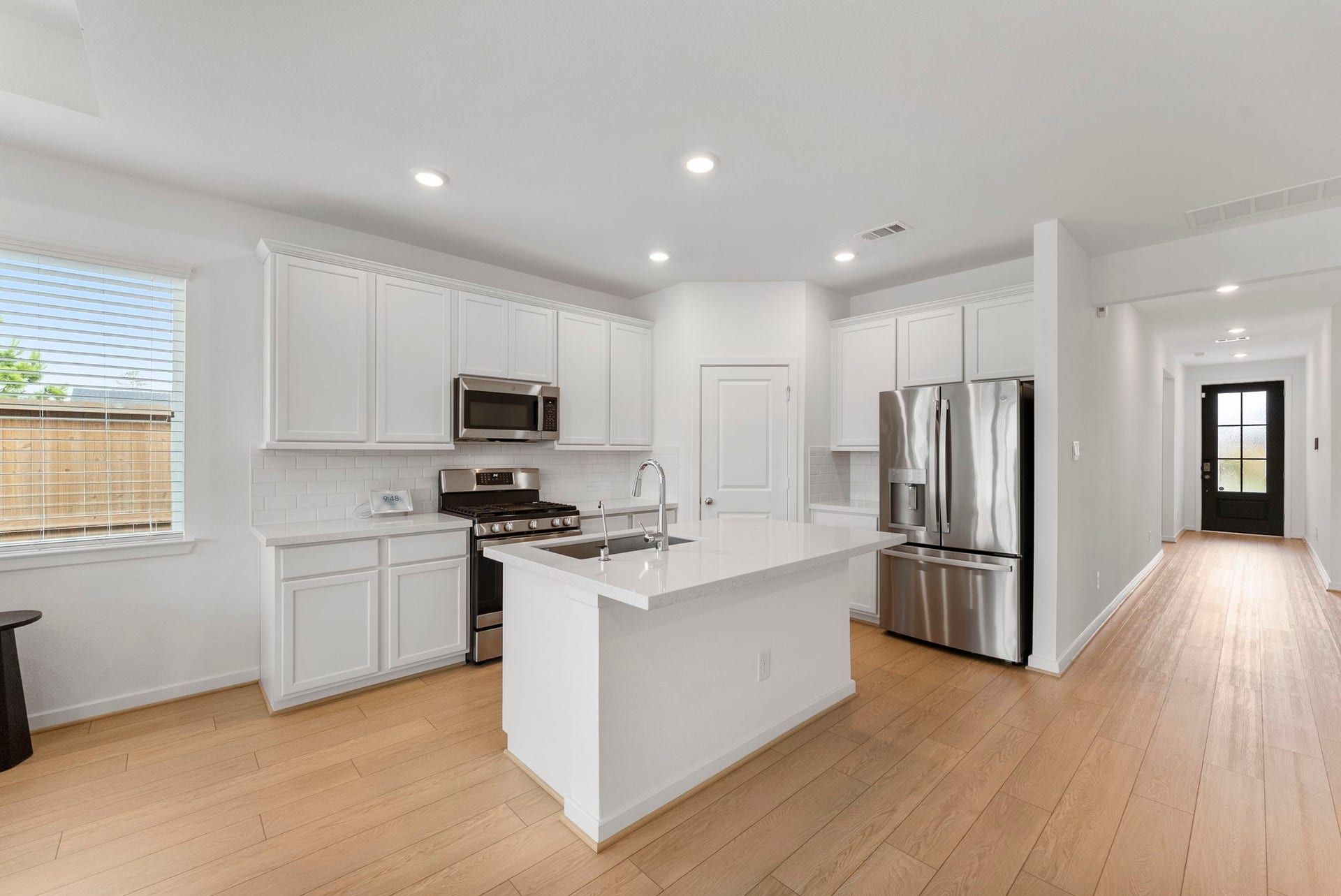 This photo showcases a modern, bright kitchen with white cabinetry, stainless steel appliances, and a central island. The open layout features wood flooring and ample natural light from a nearby window, leading to a hallway with a glass-paneled door.