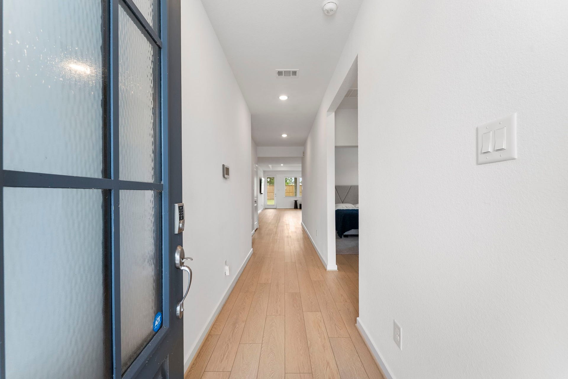 The photo shows a modern, bright entryway with light wood flooring and white walls, leading to an open living space. There's a side room visible, and recessed lighting enhances the clean, inviting atmosphere.