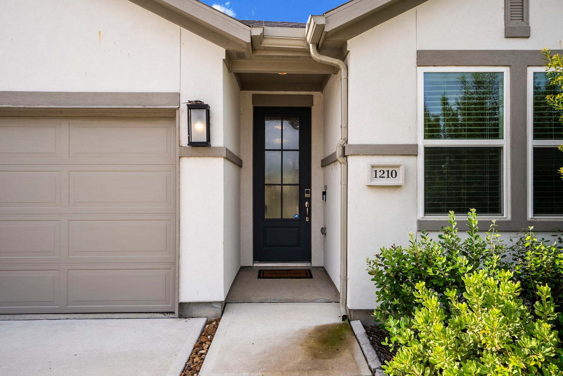 Charming home entrance with a modern black door, adjacent garage, and well-kept landscaping. The house number is clearly displayed, and the exterior features neutral tones and a welcoming ambiance.