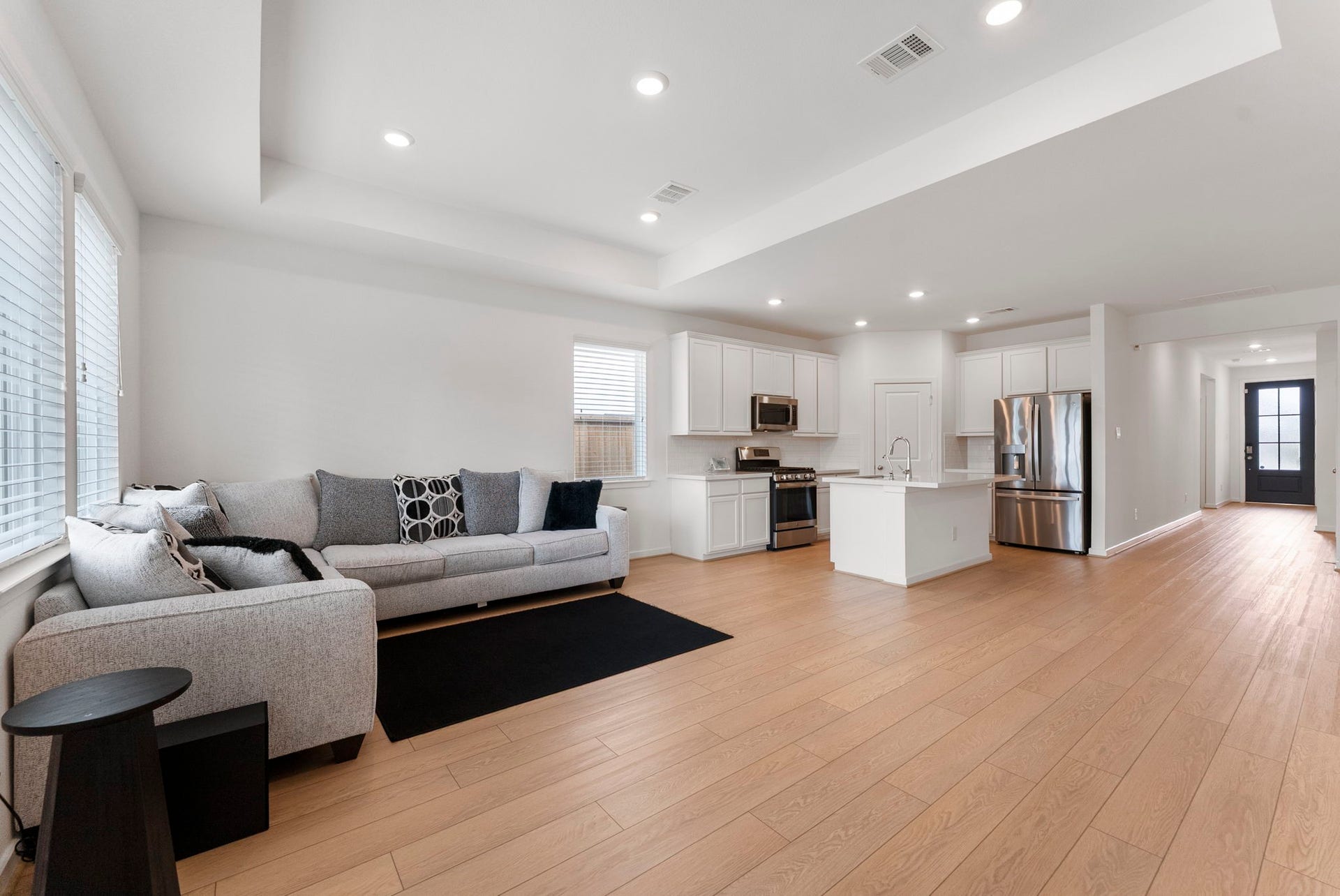 This photo showcases a modern, open-concept living space with a seamless flow between the living room and kitchen. The area features light wood flooring, a cozy gray sectional sofa, and a sleek kitchen with white cabinetry, stainless steel appliances, and a central island. Abundant natural light streams in through large windows, creating a bright and inviting atmosphere.