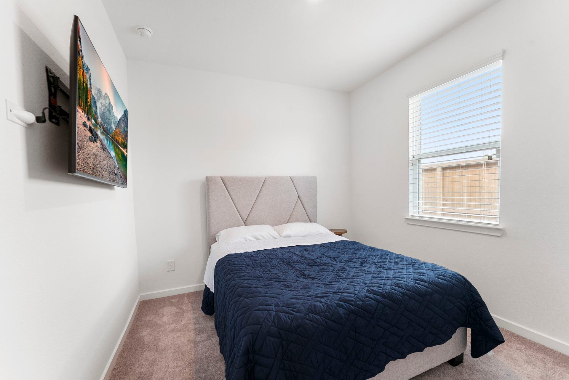 This cozy, bright bedroom features a modern bed with a blue quilt, a wall-mounted TV, and a window for natural light. Perfect for relaxation and comfort.