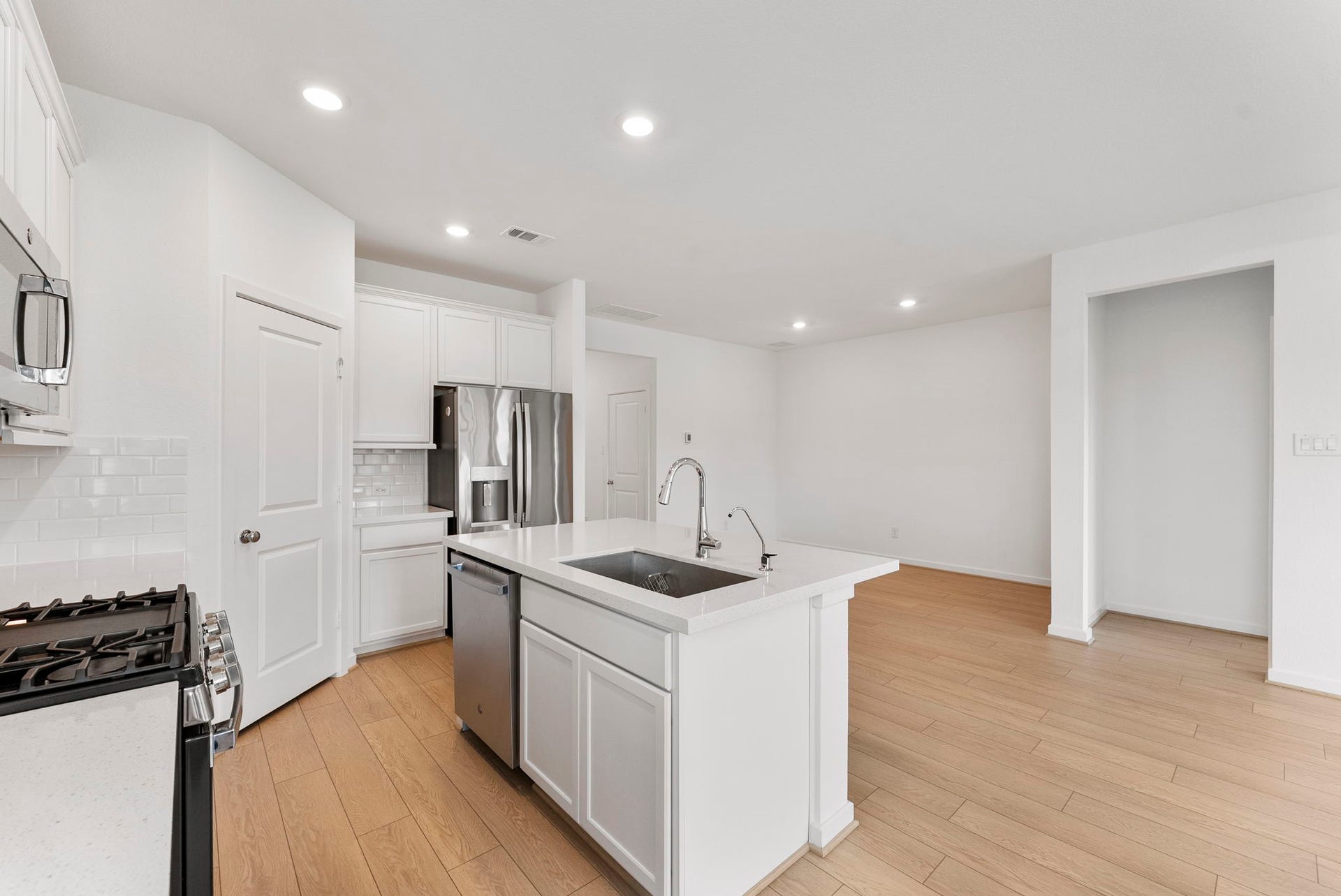 Modern kitchen with an open layout, featuring white cabinetry, stainless steel appliances, and a central island with a sink. The space is bright with recessed lighting and light wood flooring.