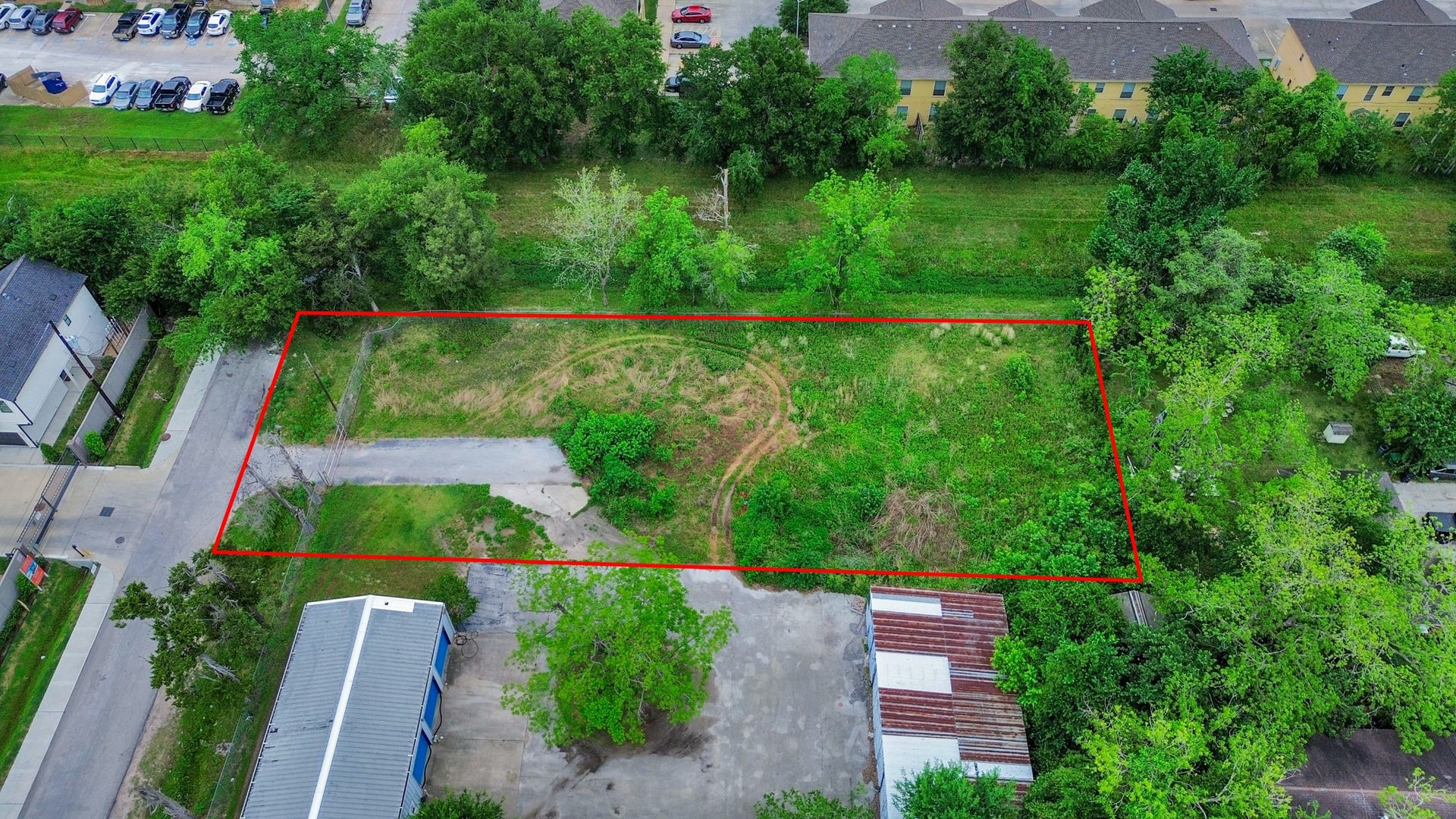 This aerial photo shows a rectangular plot of land outlined in red, featuring a mix of grass and trees. It's situated in a residential area with nearby roads and buildings, offering potential for development.