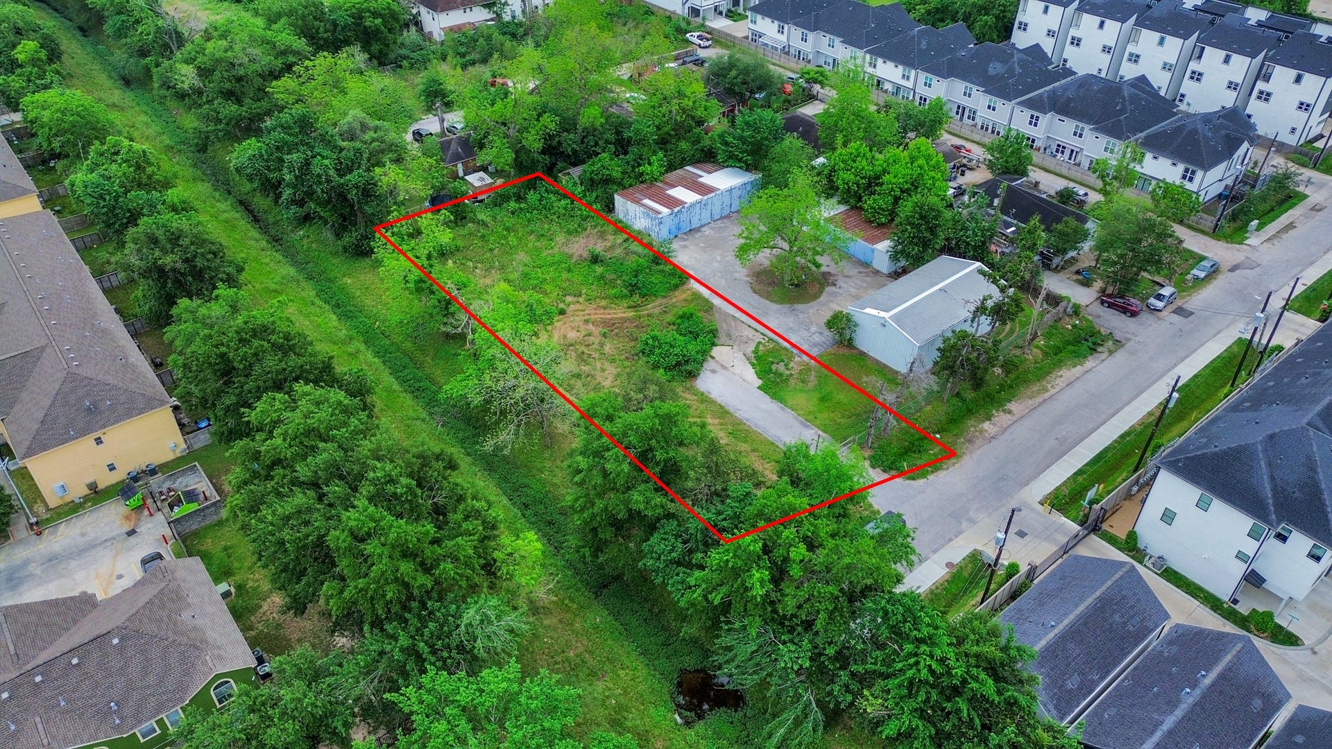This aerial photo shows a rectangular plot of land outlined in red, surrounded by greenery and residential buildings. The lot appears to have a driveway and some structures, offering potential for development in a quiet neighborhood setting.