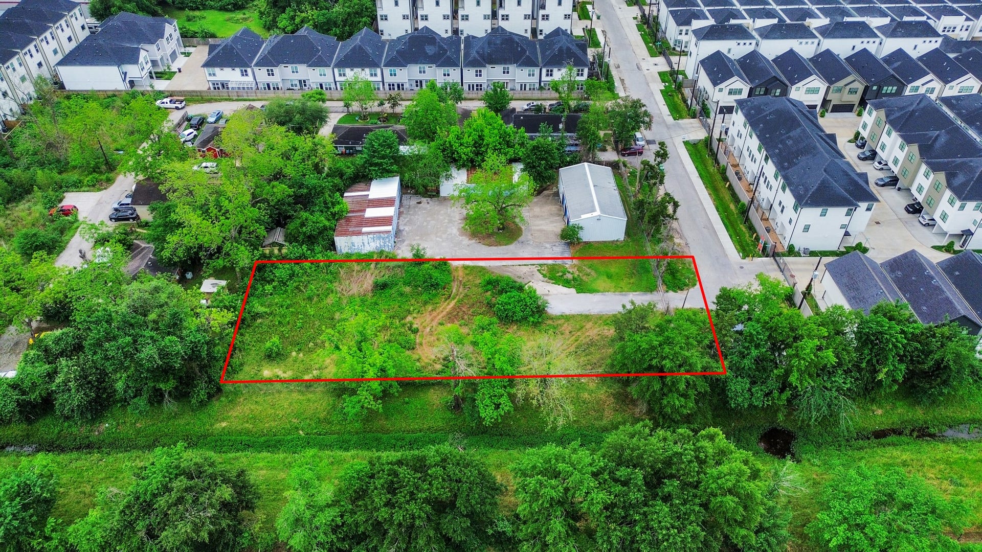 This aerial photo shows a rectangular plot of land outlined in red, surrounded by greenery. It's located in a residential neighborhood with nearby modern townhouses. The plot appears to be largely undeveloped, offering potential for building.