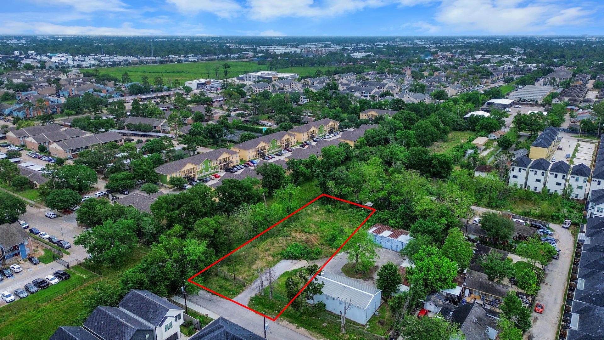 This photo shows a plot of land outlined in red, surrounded by greenery and trees in a residential neighborhood. Nearby are various houses and apartment buildings, providing a suburban setting. The area appears peaceful with ample space and potential for development.