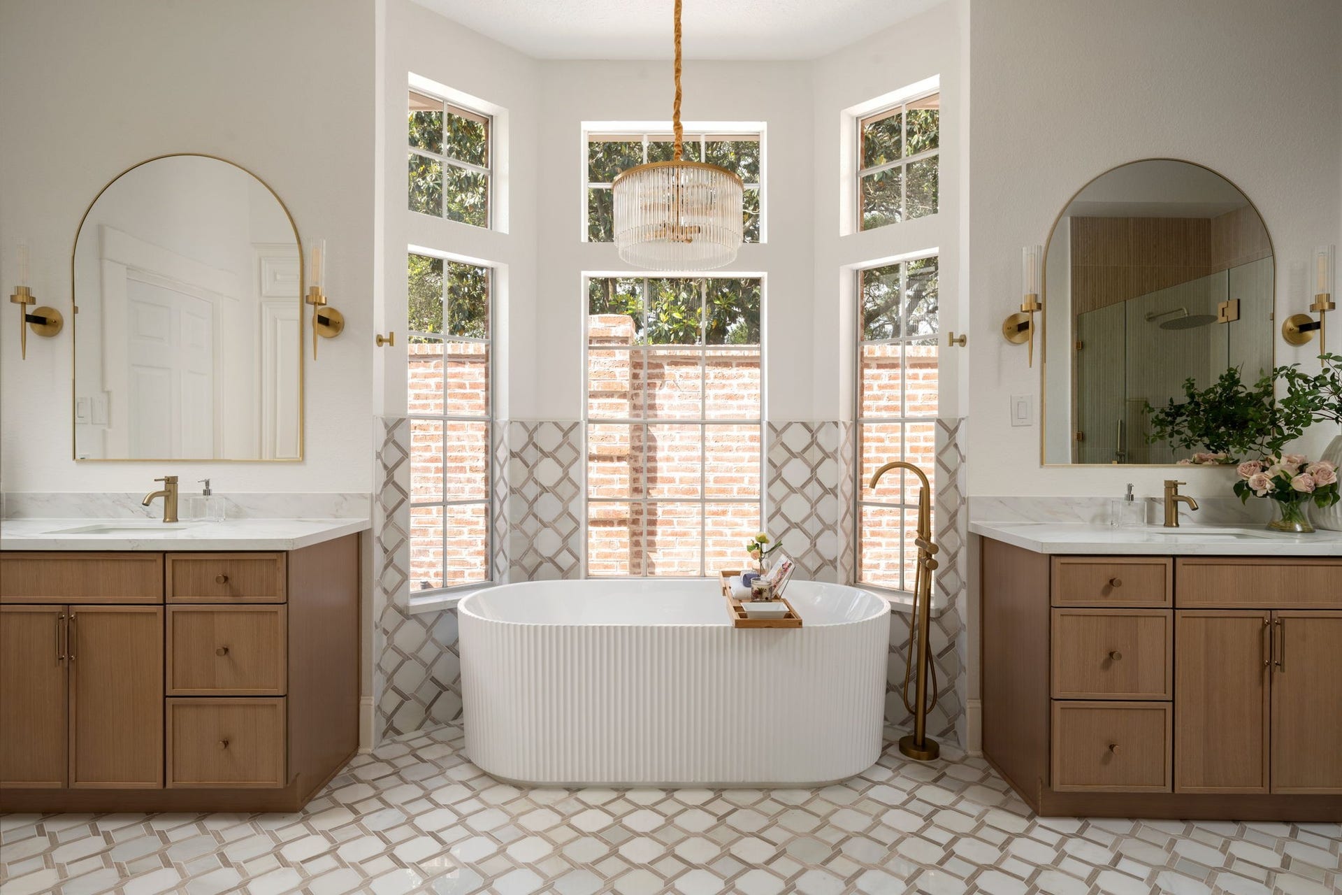 The primary bath features a freestanding soaking tub positioned beneath oversized windows and surrounded by custom marble flooring and tilework. Dual vanities, brass fixtures, and contemporary lighting contribute to the spaces spa-inspired design. Natural light fills the room throughout the day while maintaining privacy.