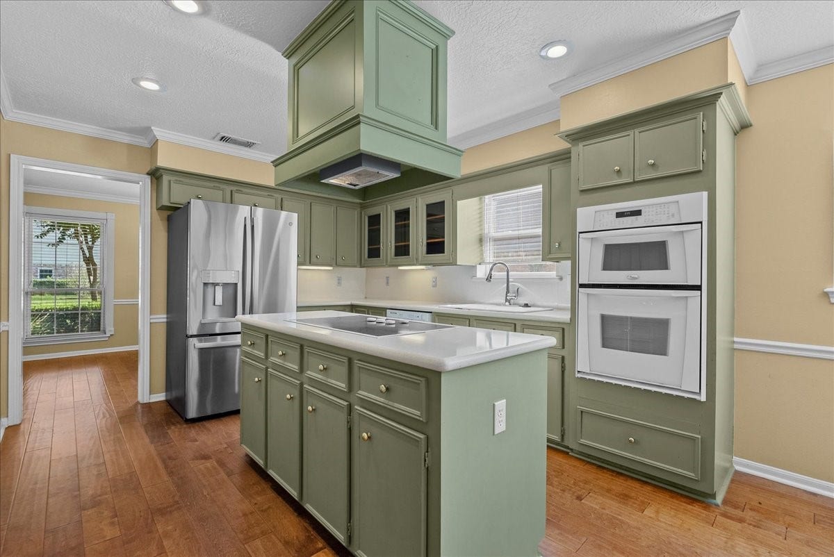 Charming kitchen with sage green cabinetry, sleek stainless steel appliances, and a central island with cooktop. Natural light streams through the window, highlighting the warm wood flooring and classic double oven setup.