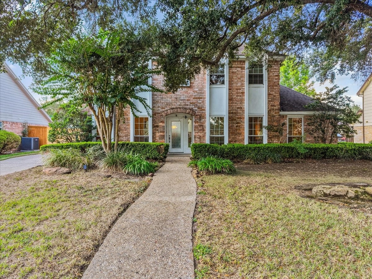 Charming two-story brick home with a welcoming pathway, surrounded by lush greenery and mature trees.