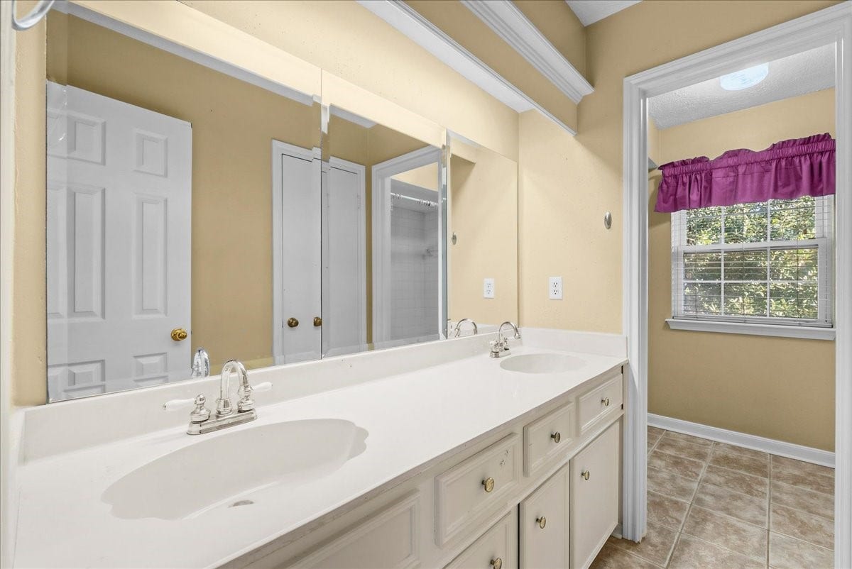 Spacious bathroom featuring a dual-sink vanity, elegant light fixtures, and a large mirror, complemented by natural light from the window with a charming purple valance.