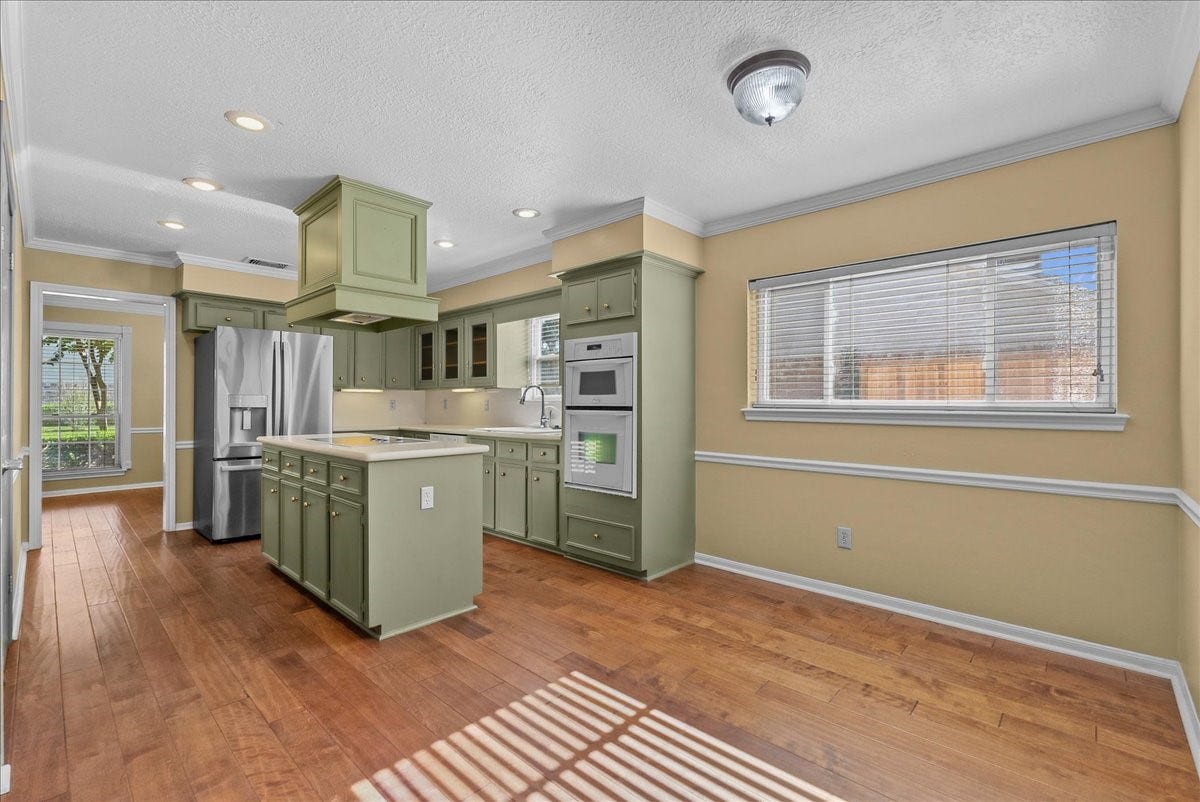 Spacious kitchen with charming sage green cabinetry, an island with a cooktop, and stainless steel appliances, complemented by warm wood flooring and abundant natural light.