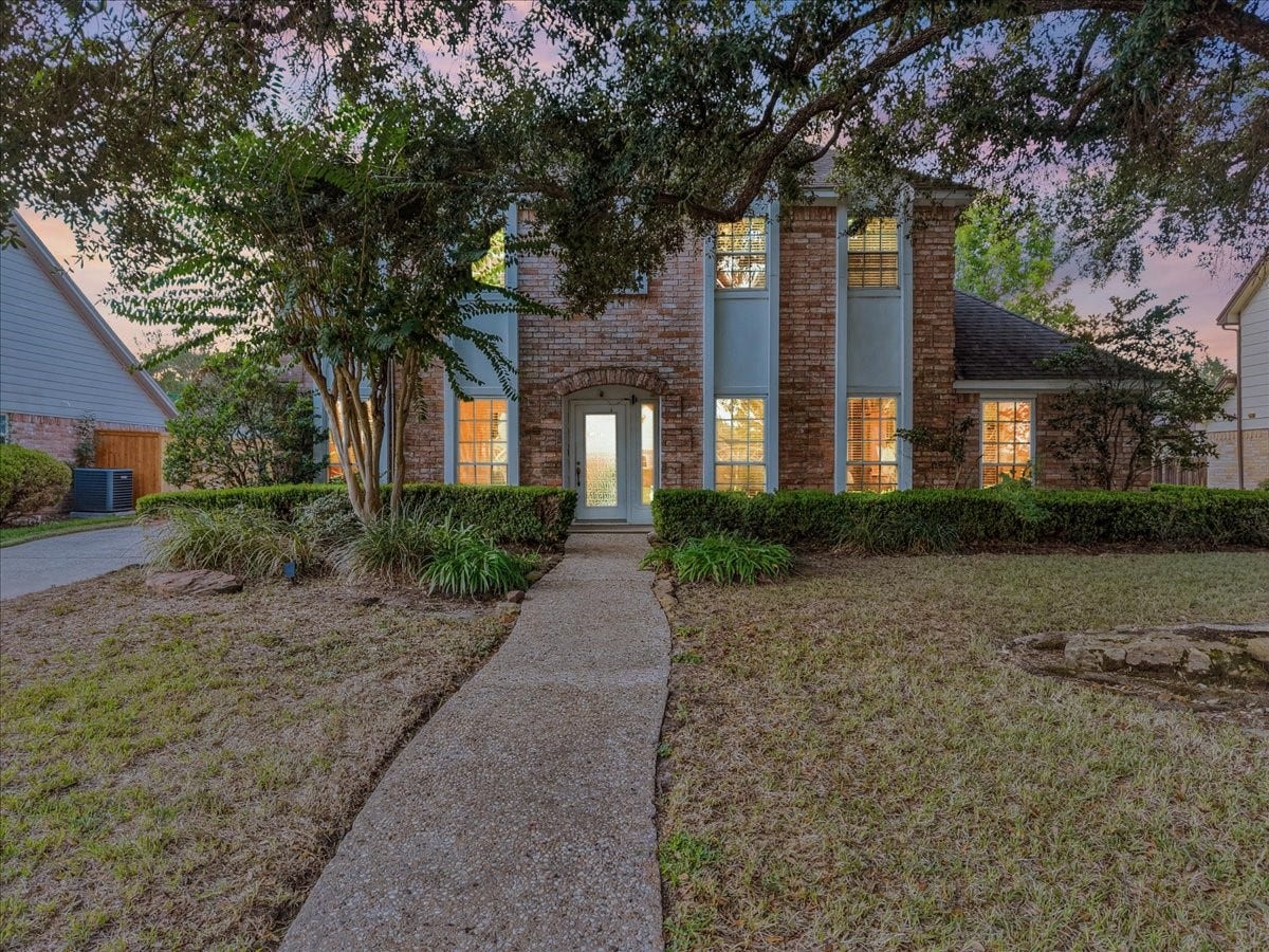 Charming two-story brick home with elegant columns, surrounded by lush greenery and mature trees, offering a warm and inviting entrance.