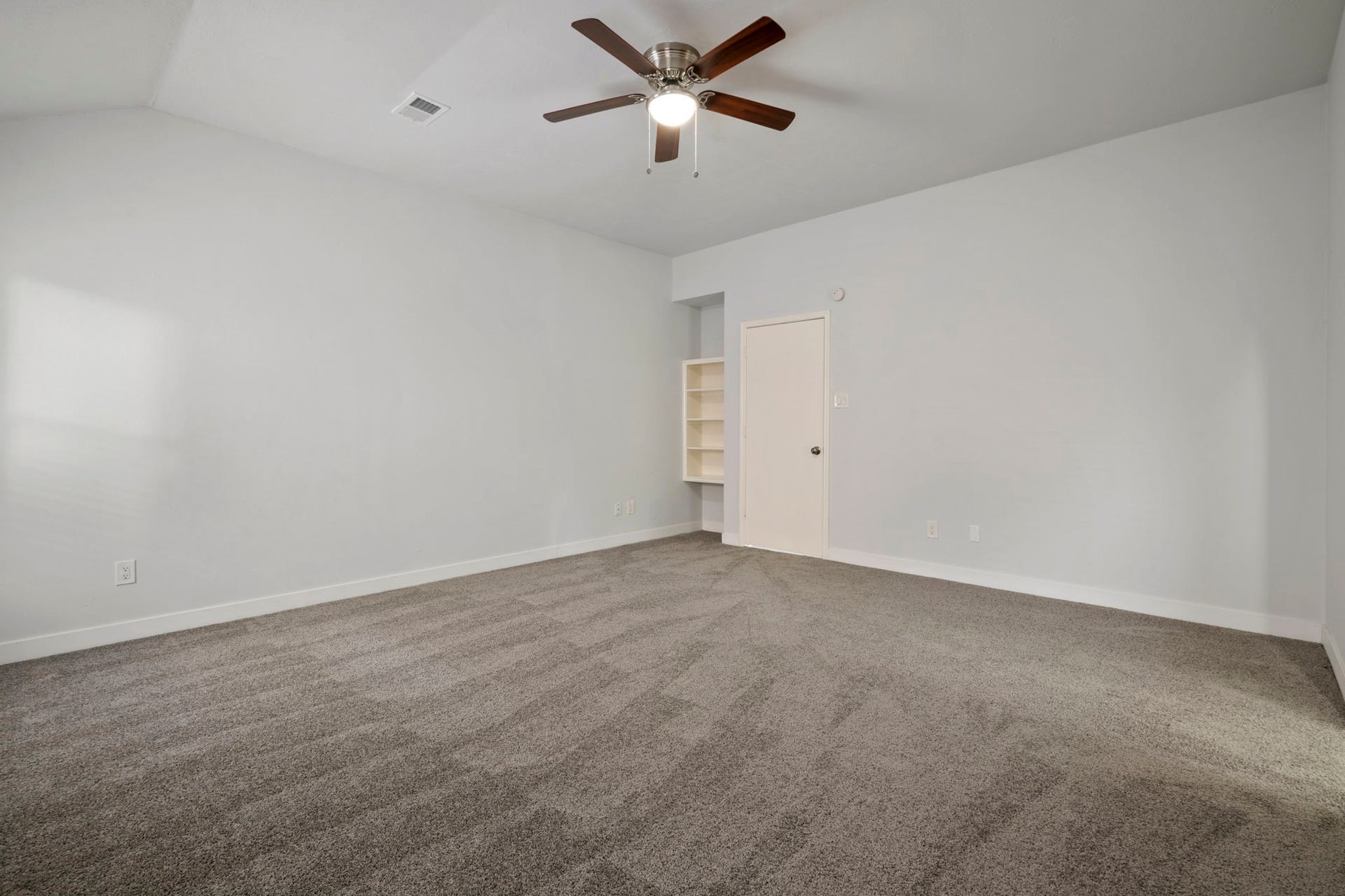 Spacious bedroom with soft carpet, neutral walls, and a ceiling fan, featuring built-in shelving and ample natural light.