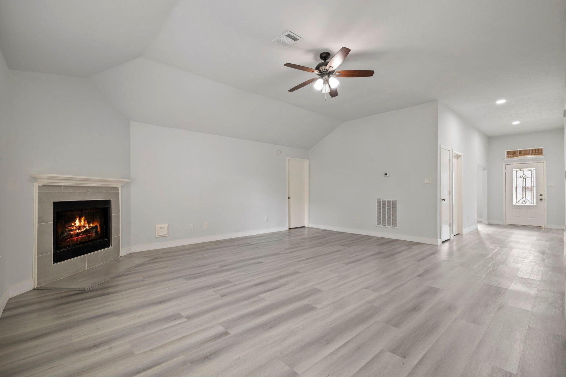 Spacious living room with sleek hardwood floors, a cozy corner fireplace, and a ceiling fan, leading to a bright entryway.